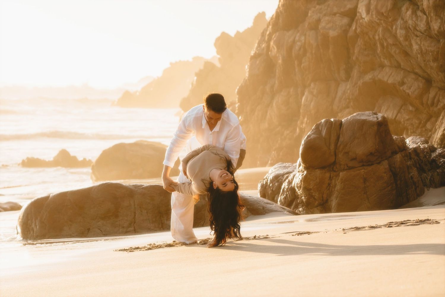 Couple among beach boulders as he dips her backwards, seen from a distance