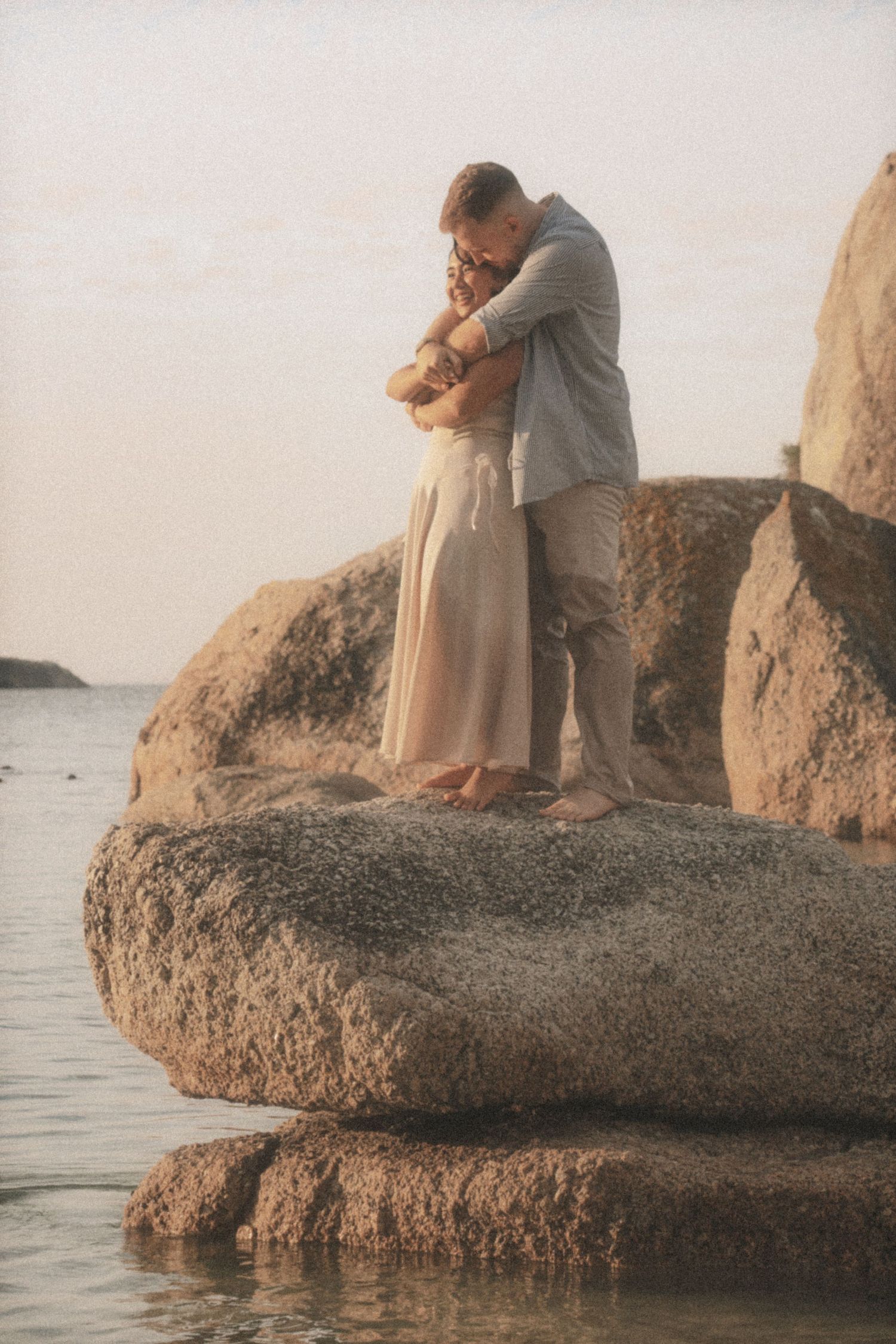 Couple embracing on top of a large boulder with the sea behind them