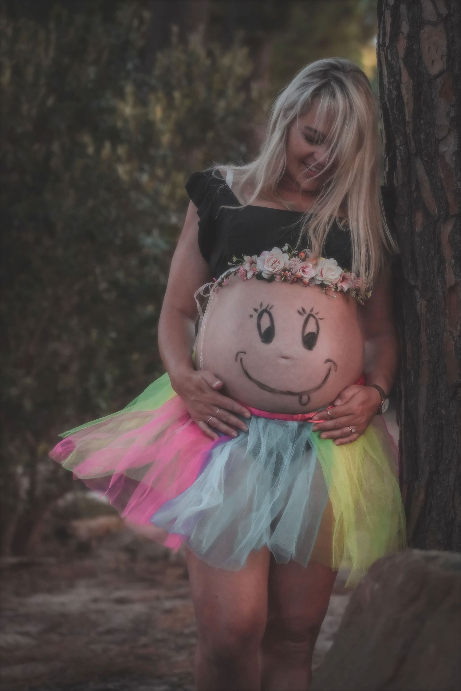 Pregnant woman with a smiley face painted on her bump, wearing a flower crown and rainbow tutu