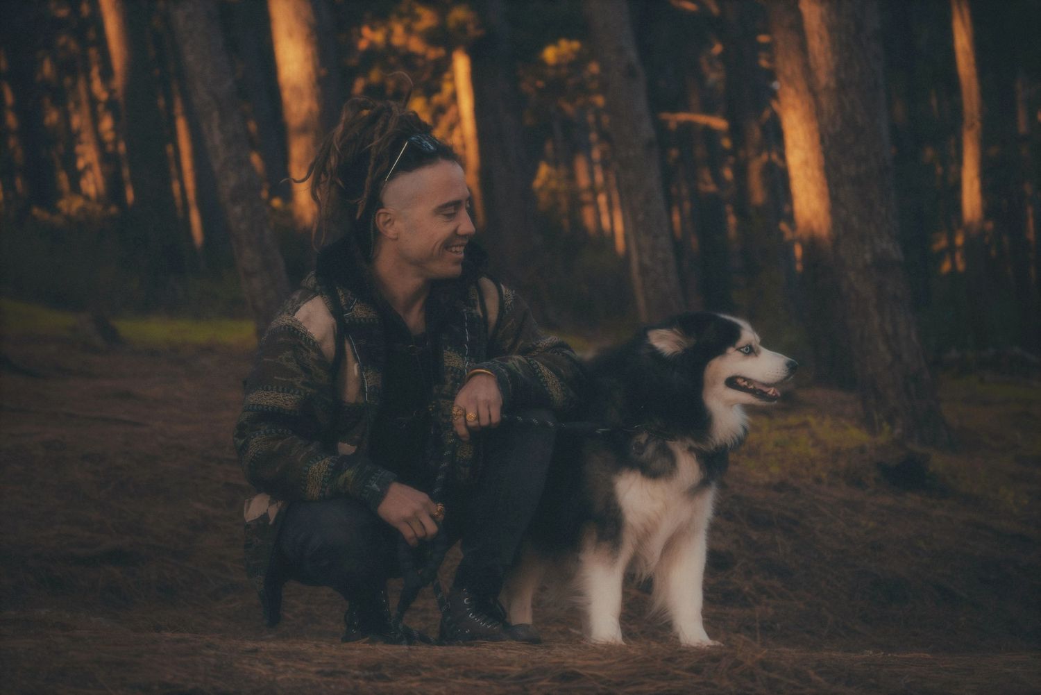 Man crouching with his husky between the pines at golden hour