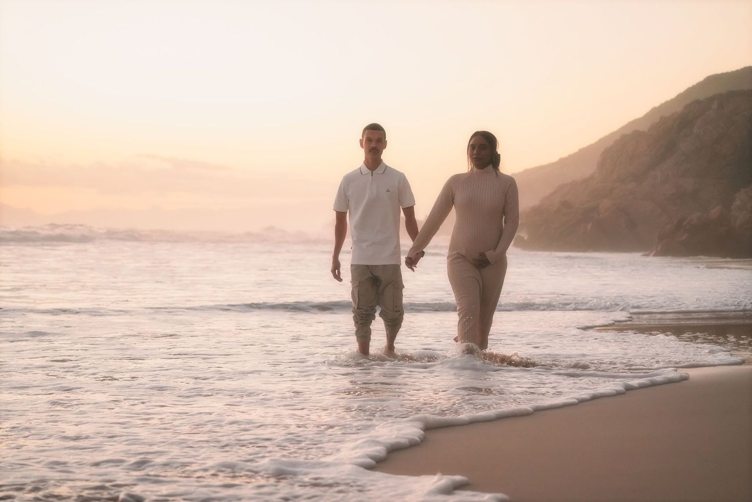 Couple walking hand in hand through shallow waves with mountains in the background