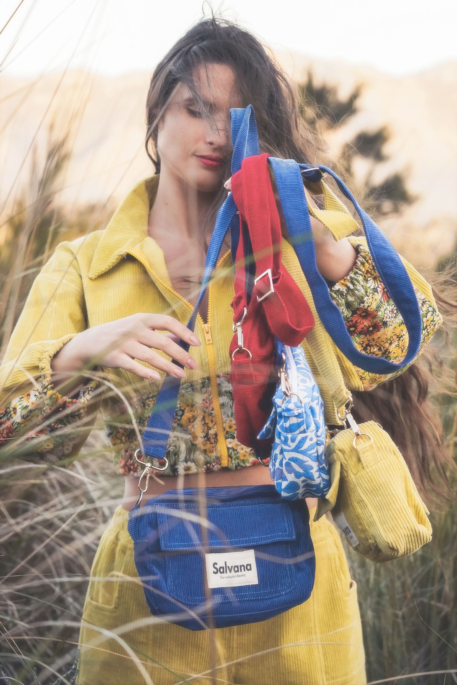 Woman in a yellow jacket holding red and blue corduroy bags up to her face