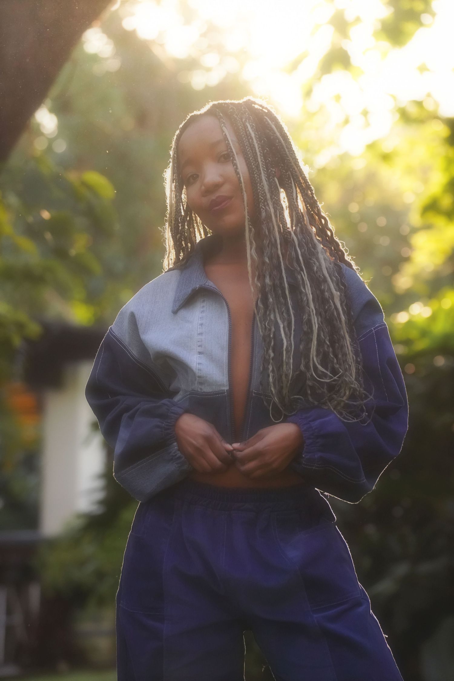 Woman with long braids in a blue and purple outfit, backlit by golden light in a garden