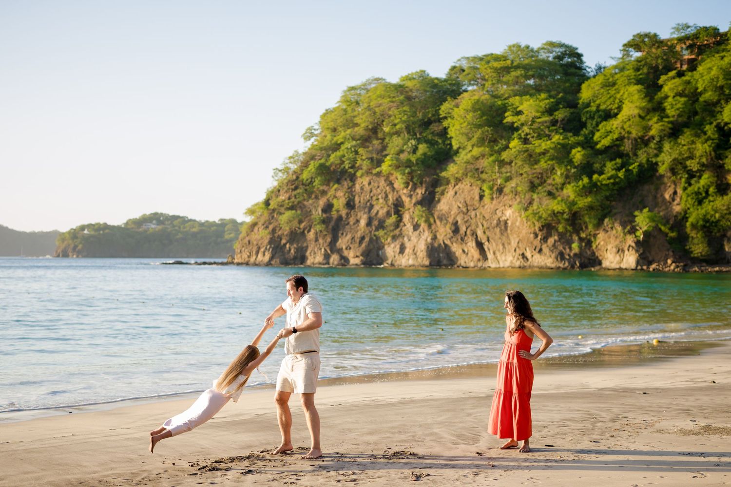 Family playing on a tropical beach with turquoise water and lush green cliffs in the background.