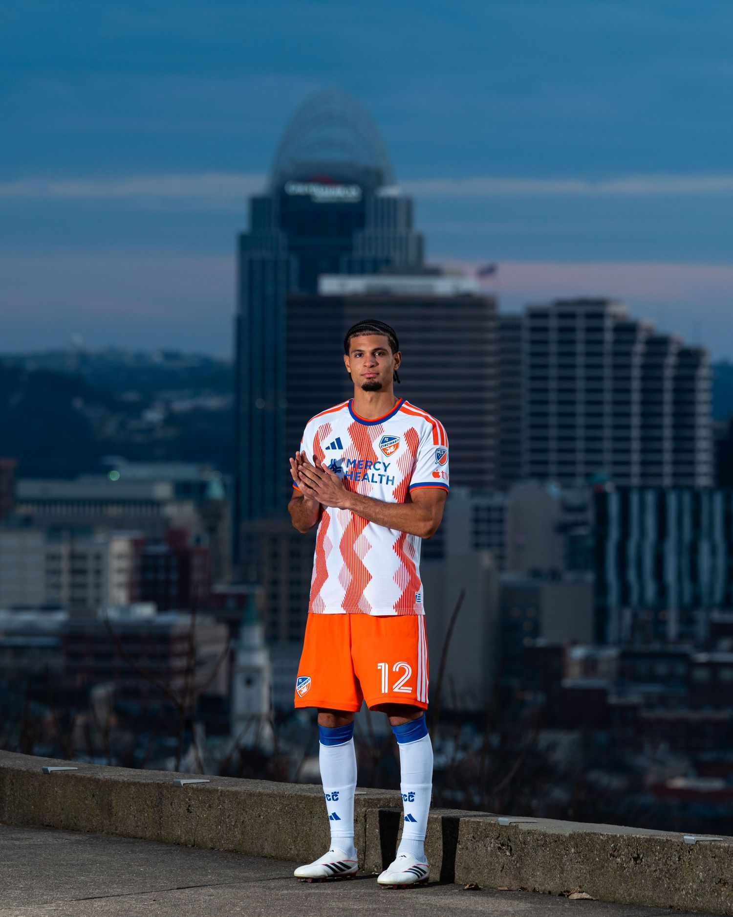Person wearing a white and orange FC Cincinnati uniform standing on an overlook with the Cincinnati skyline in the background at dusk.