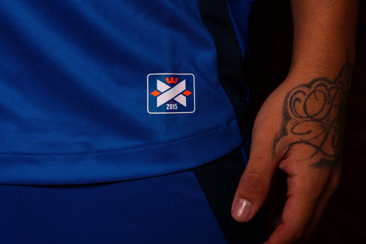 Close-up detail of the lower portion of a blue FC Cincinnati uniform showing a commemorative crest patch and a tattooed hand.