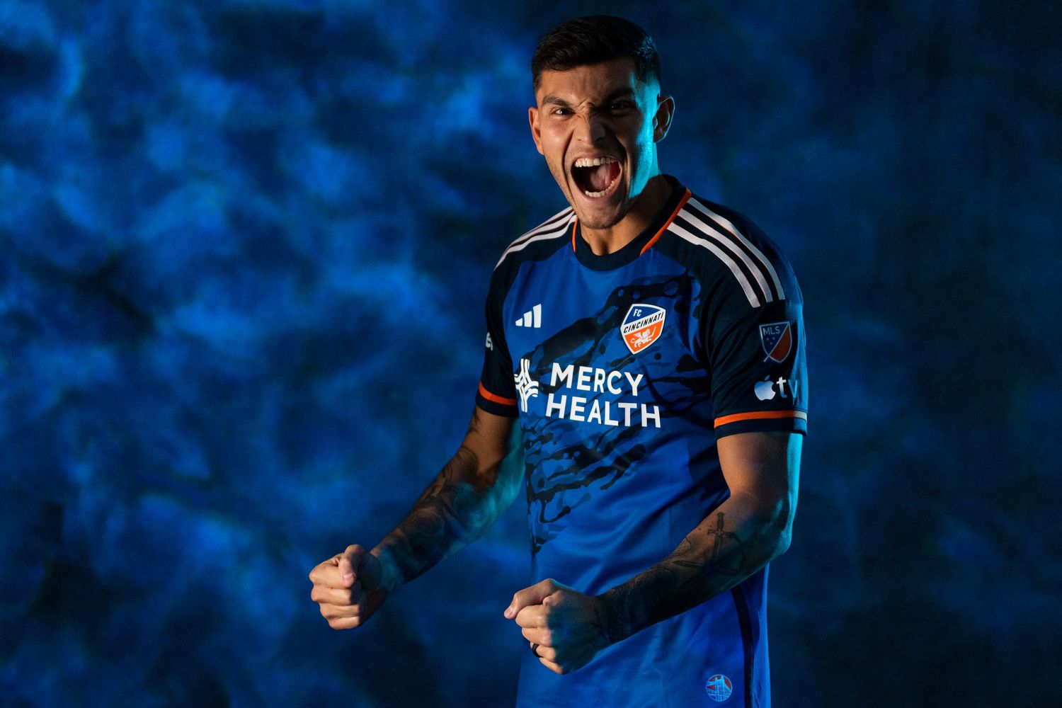 Person wearing a blue FC Cincinnati jersey standing against a blue textured backdrop with both hands clenched in front of the body.