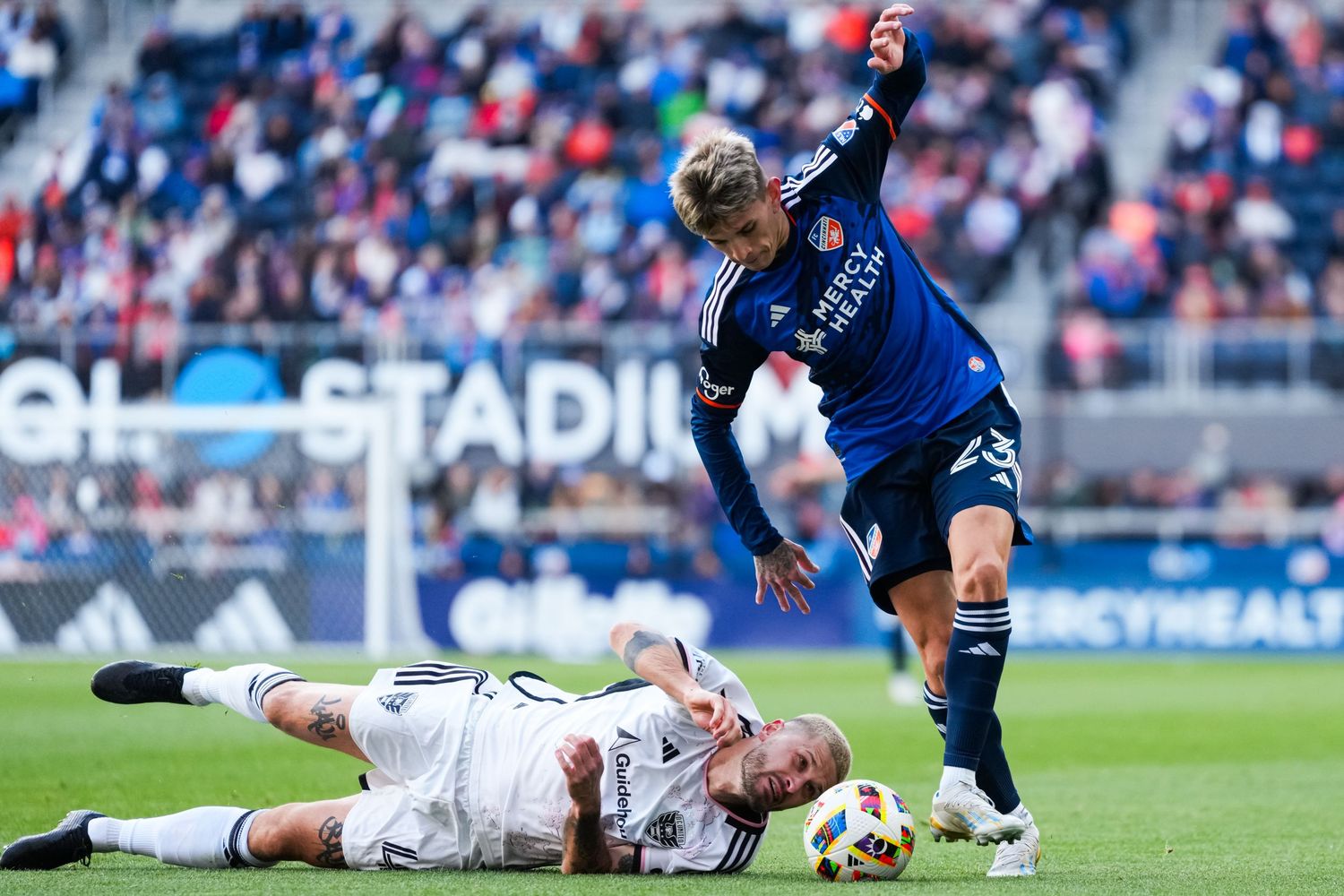Soccer player challenges for possession as an opposing player lies on the field next to the ball.