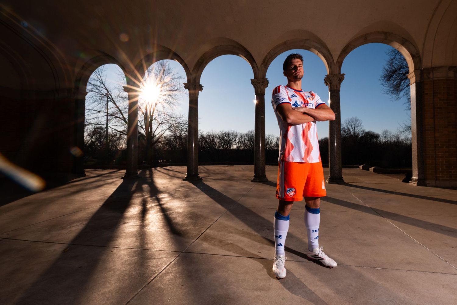 Person wearing a white and orange FC Cincinnati uniform standing with arms crossed beneath a row of stone arches, with sunlight.