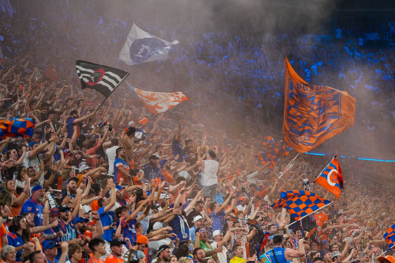FC Cincinnati supporters wave flags and cheer in a packed stadium section filled with smoke and team colors.