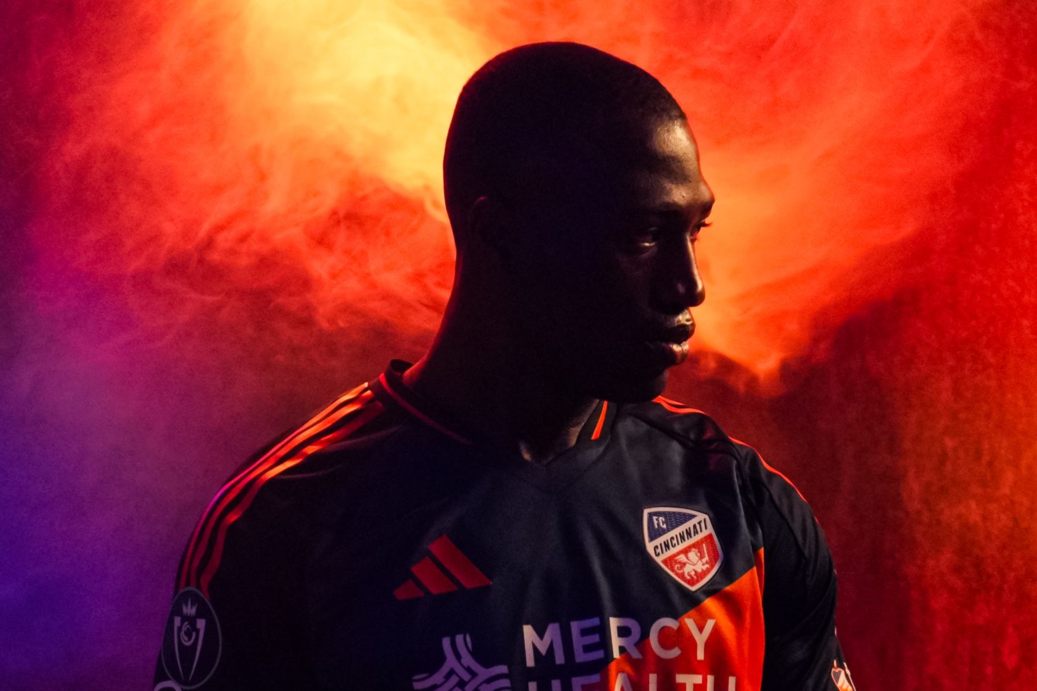 Soccer player wearing a dark FC Cincinnati jersey stands in dramatic colored lighting and smoke.