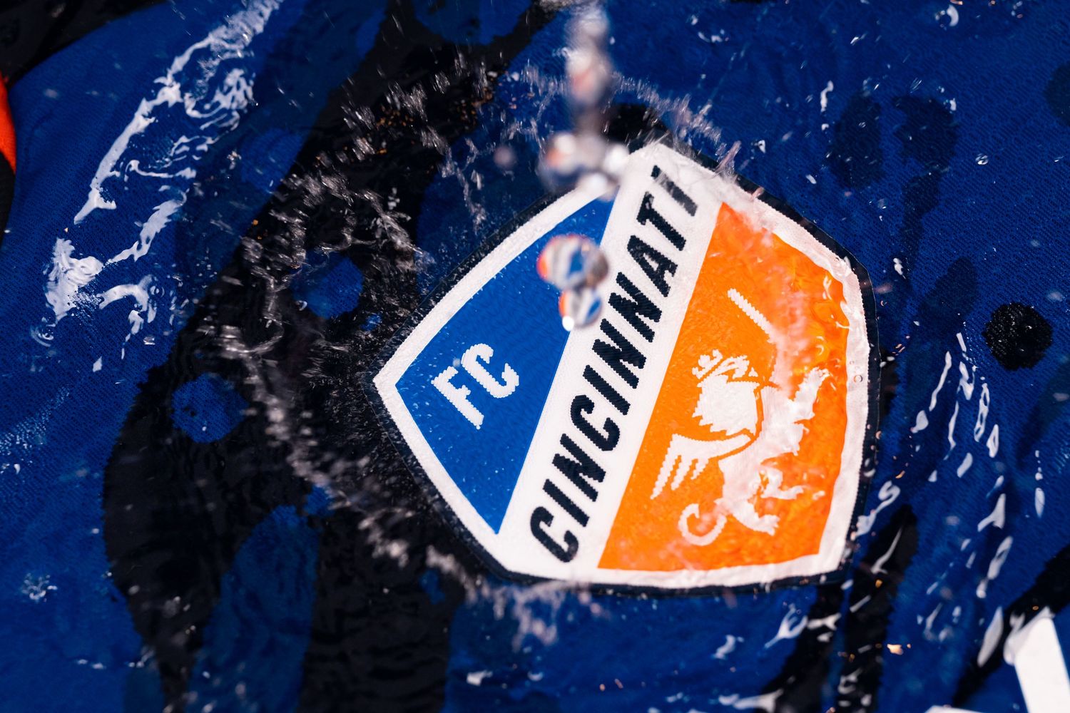 Close-up of the FC Cincinnati crest on a blue jersey as water splashes across the surface of the fabric.