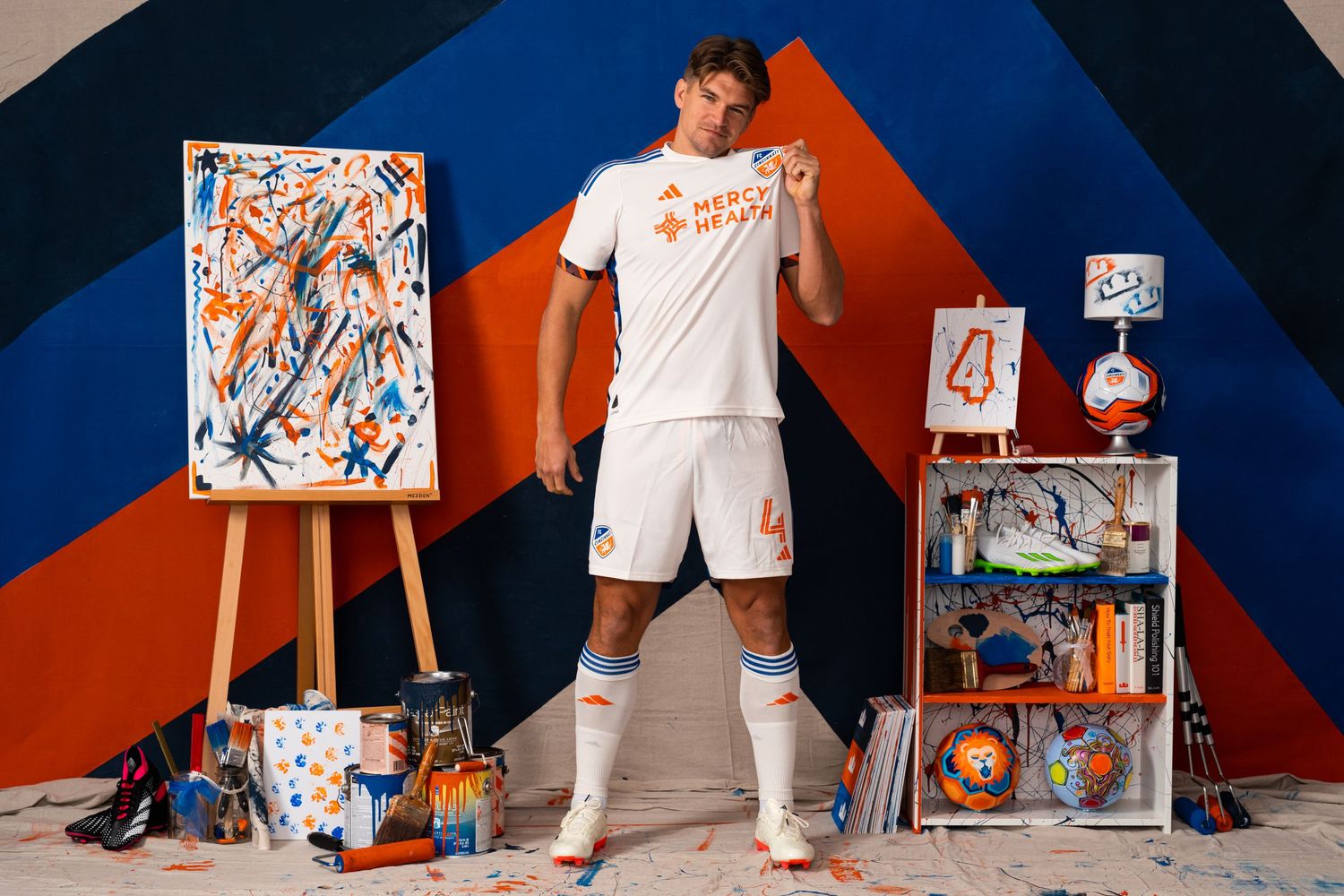 Wide studio portrait of a soccer player in a white FC Cincinnati uniform standing among paint supplies, artwork, and soccer-themed props.