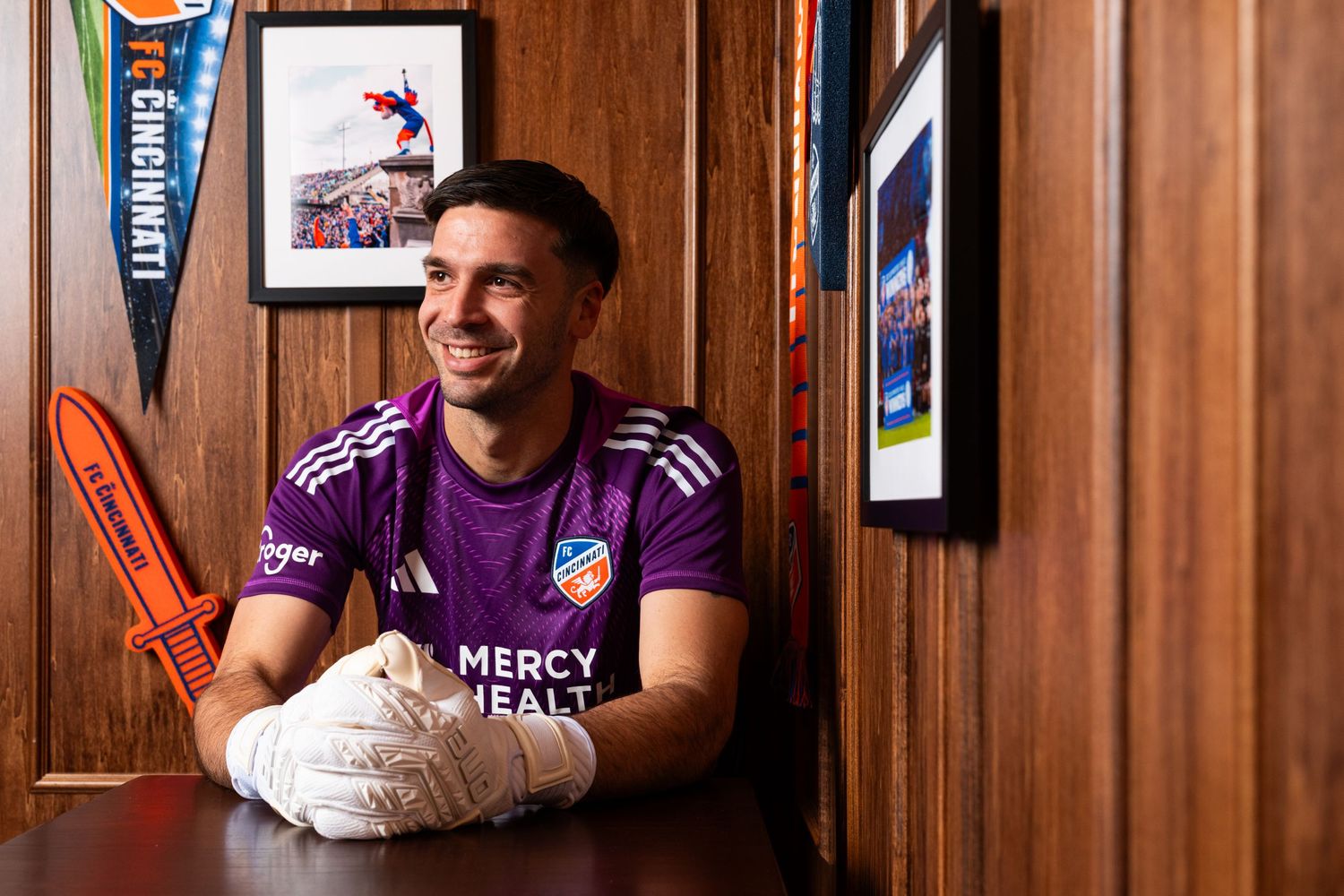 Person wearing a purple FC Cincinnati goalkeeper jersey and white goalkeeper gloves seated at a table in a wood-paneled room.