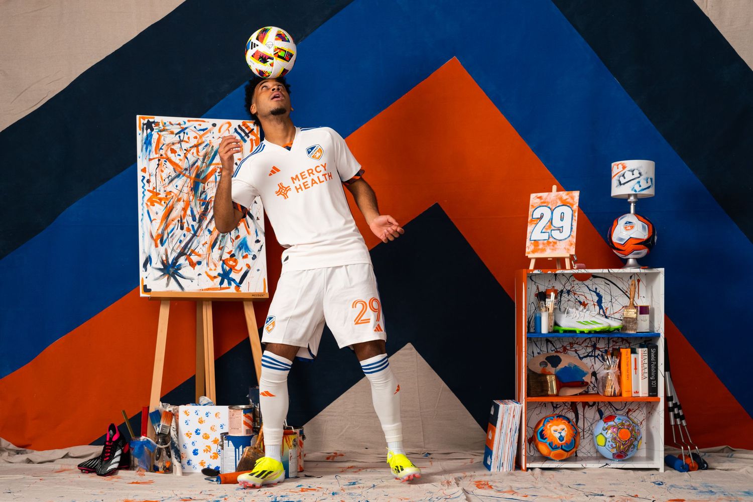 Soccer player balancing a soccer ball above their head while standing in an art-themed studio with painted canvases and FC Cincinnati décor.