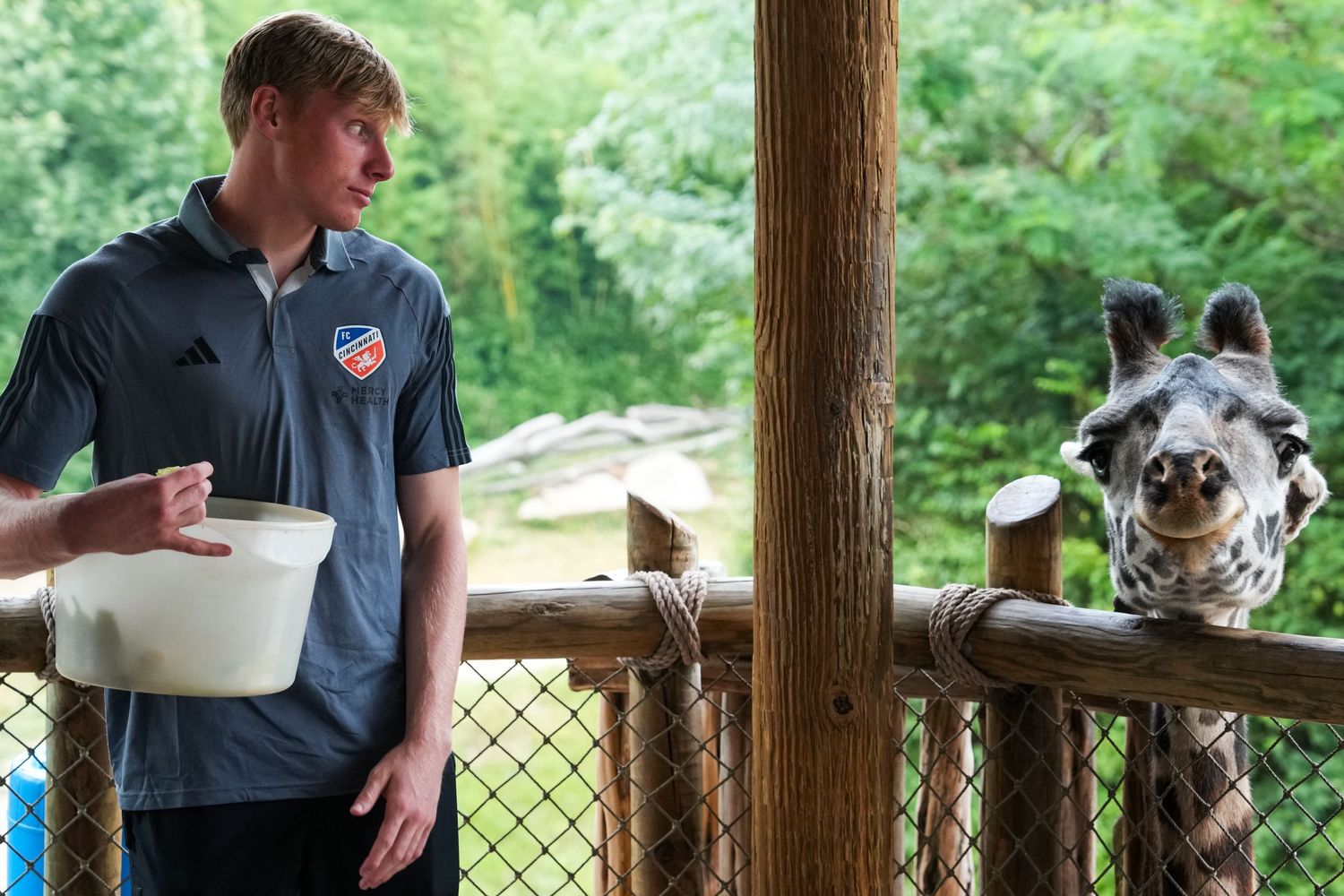 FC Cincinnati player holding a feeding bucket while standing across from a giraffe.
