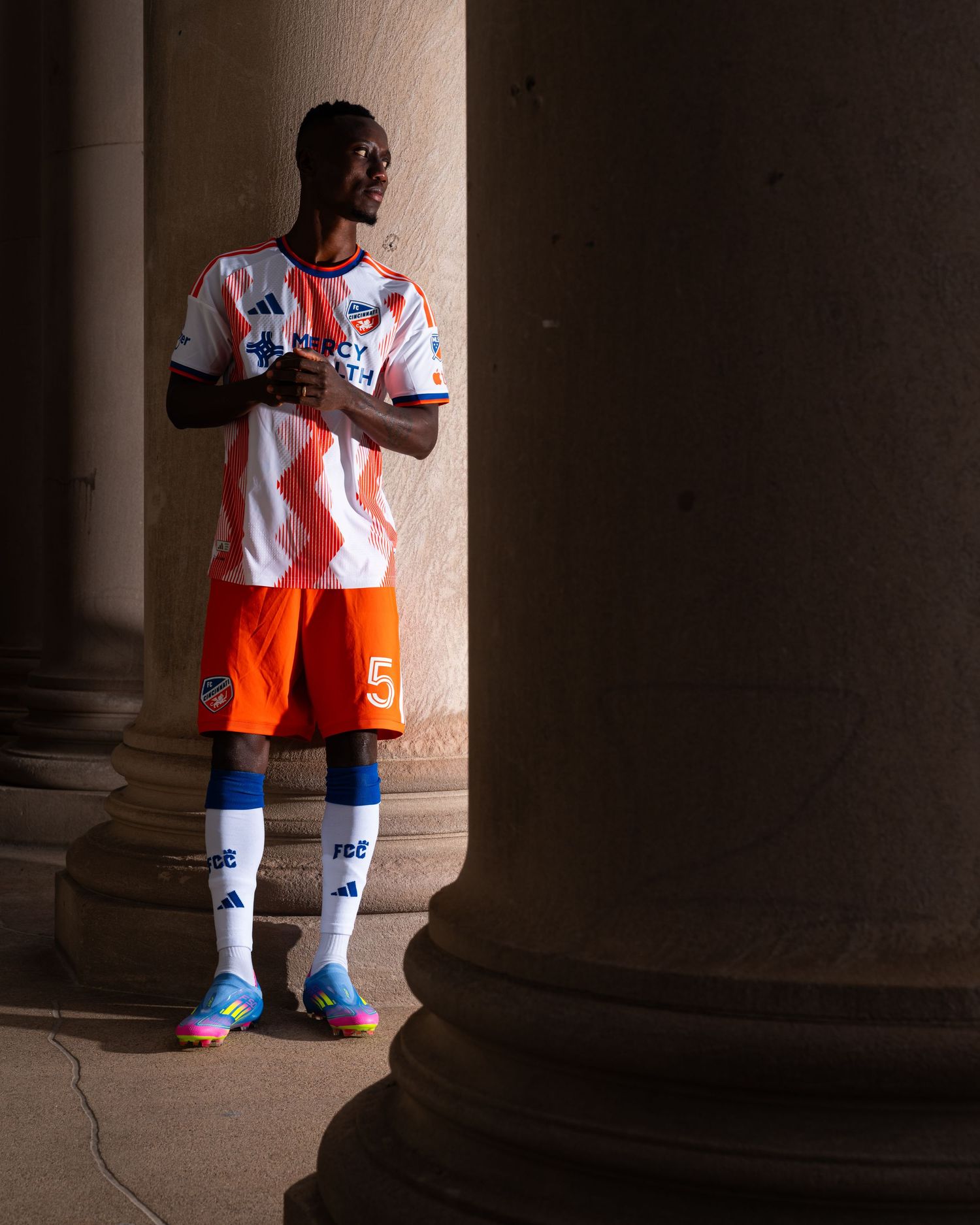 Person wearing a white and orange FC Cincinnati uniform standing between large stone columns in a historic architectural setting.
