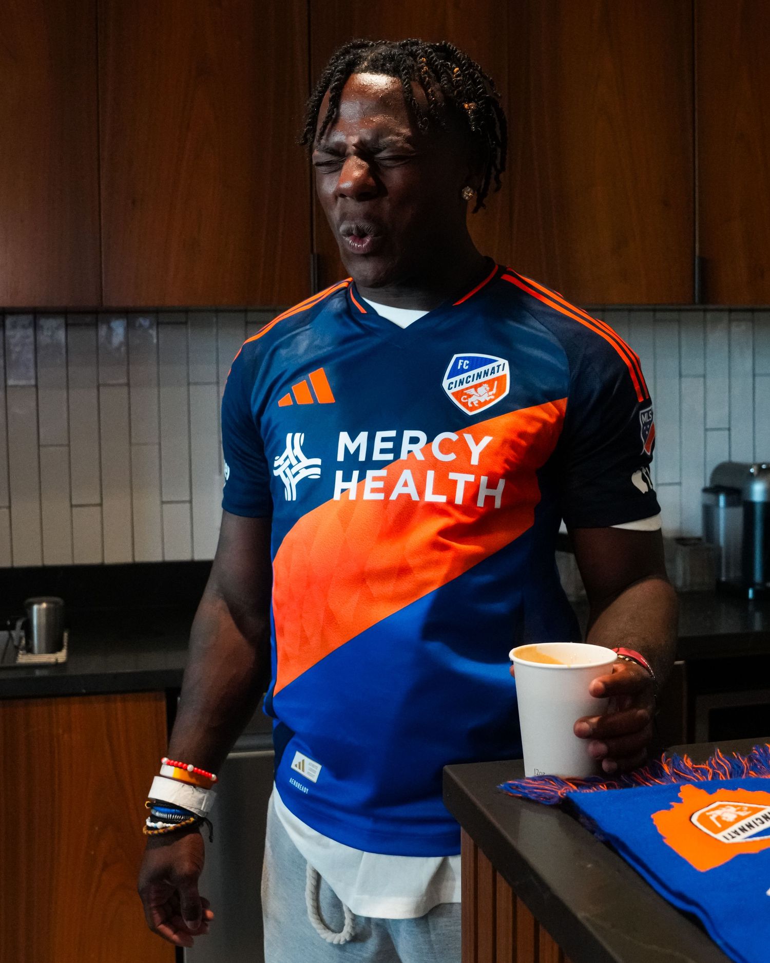 Person wearing an FC Cincinnati jersey holds a cup while standing in a kitchen setting.