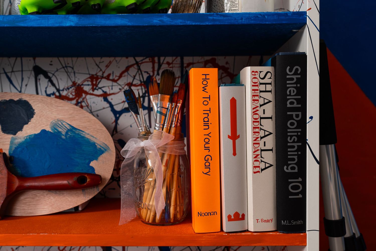 Close-up of a painted shelf displaying paintbrushes in a glass jar, an artist’s palette with blue paint, books, and art supplies.