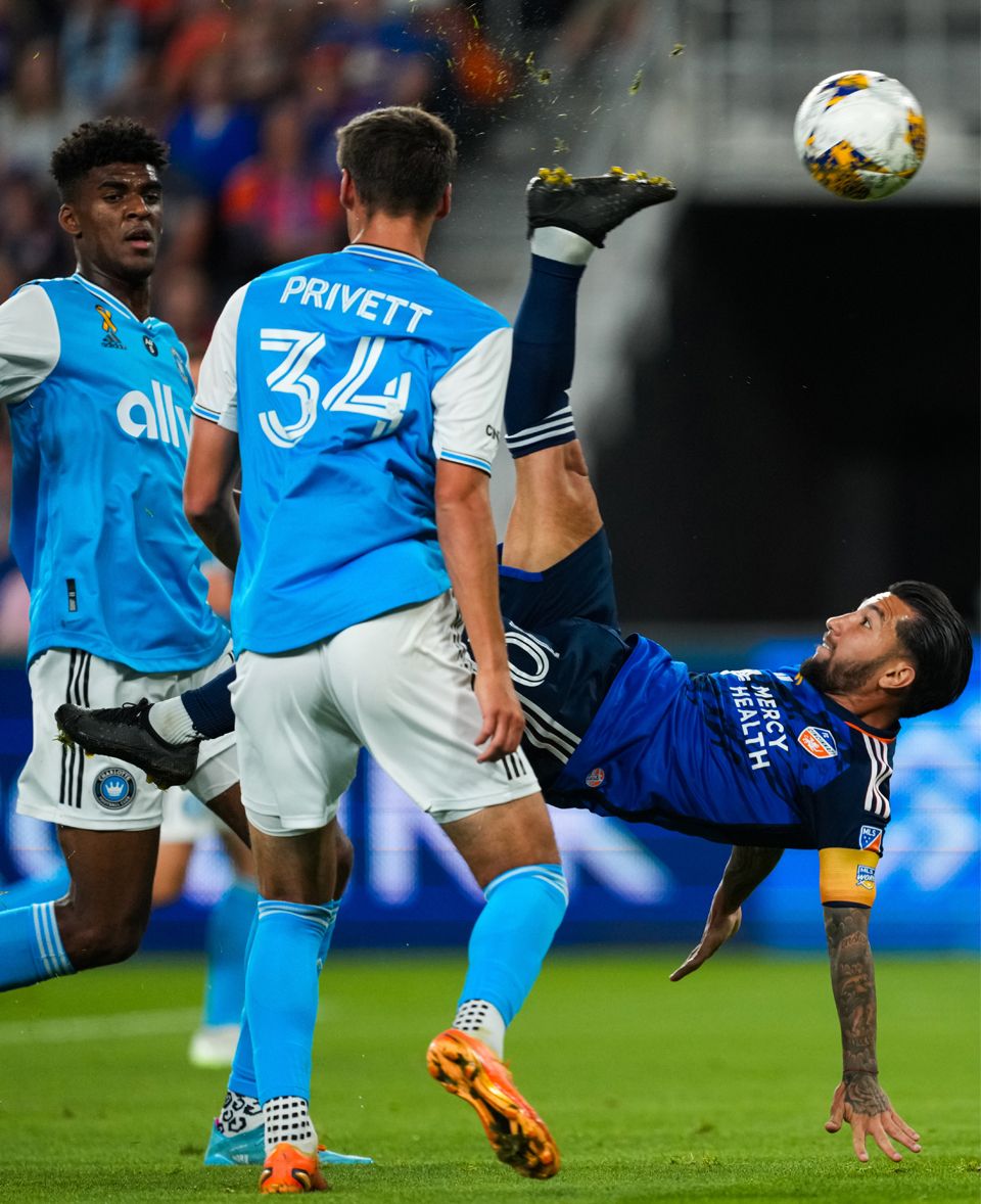 FC Cincinnati player attempts a bicycle kick while defenders watch during a soccer match.