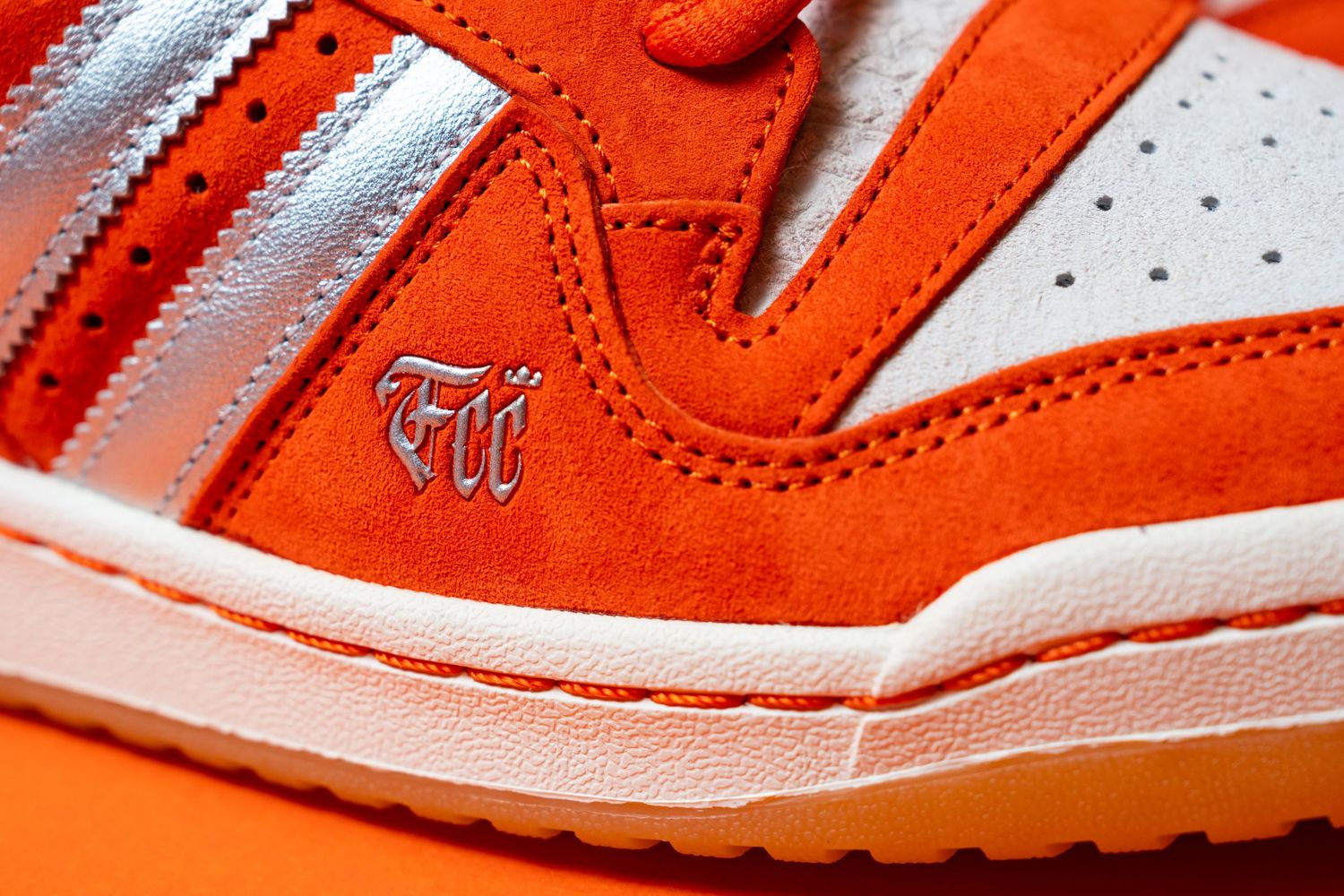 Detailed close-up of an orange suede Adidas sneaker featuring metallic silver stripes, a white toe panel, and embroidered lettering.