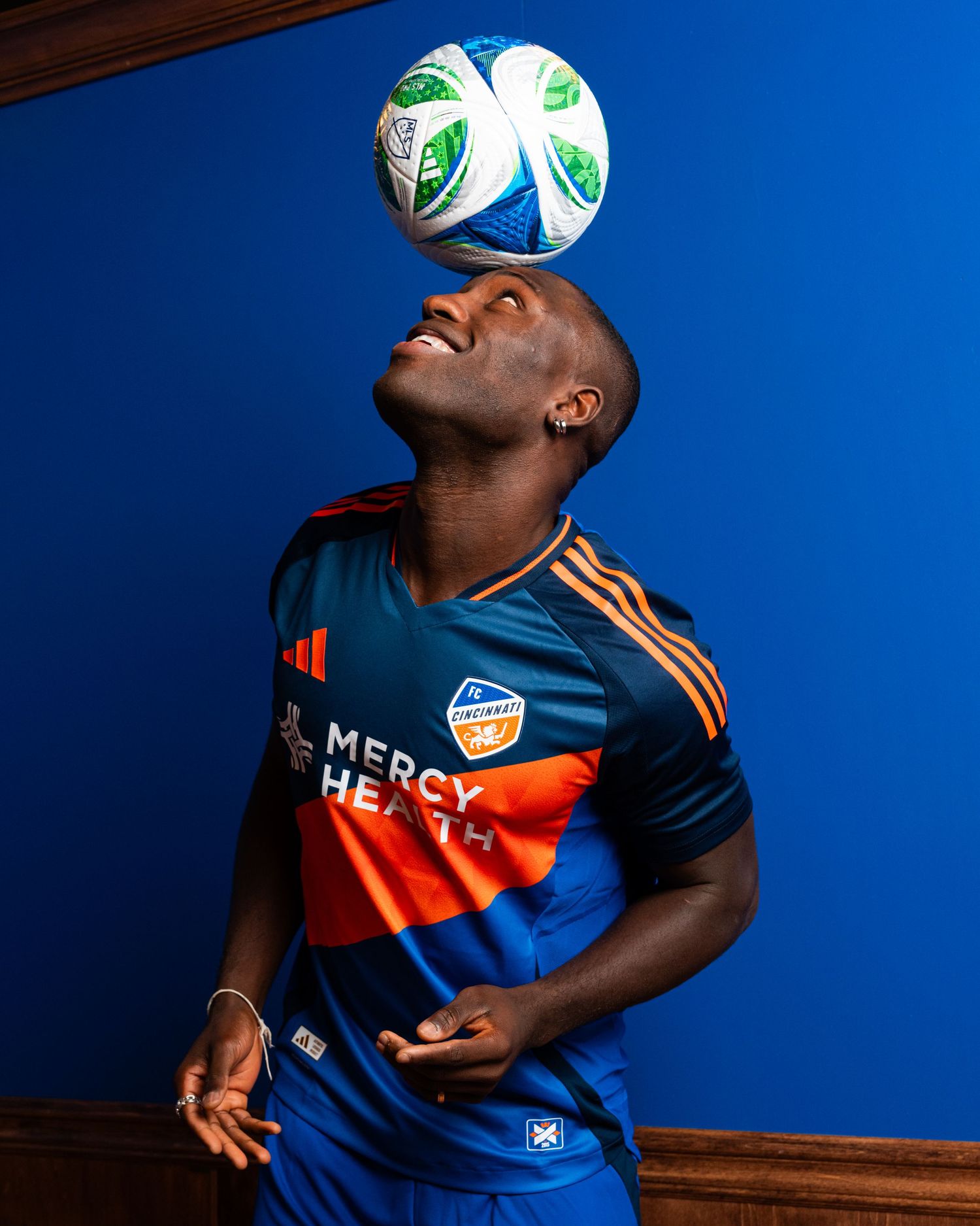 Person balancing a soccer ball on their forehead while wearing a blue and orange FC Cincinnati uniform against a blue background.
