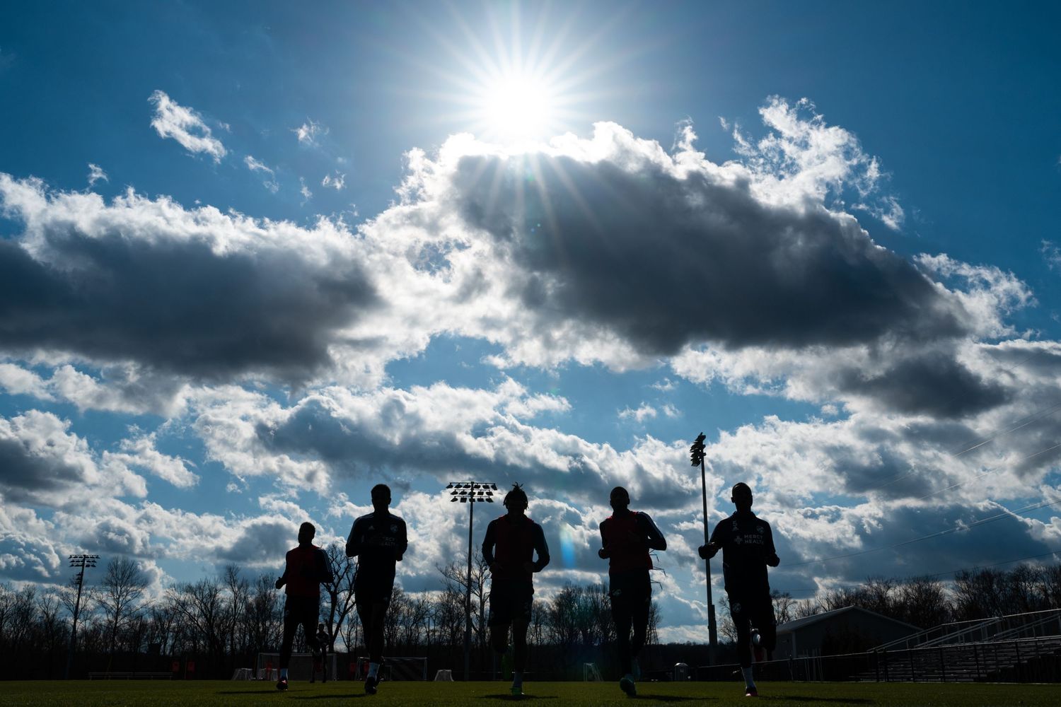 Soccer players jogging during preseason training beneath a dramatic sky with sunlight breaking through clouds.