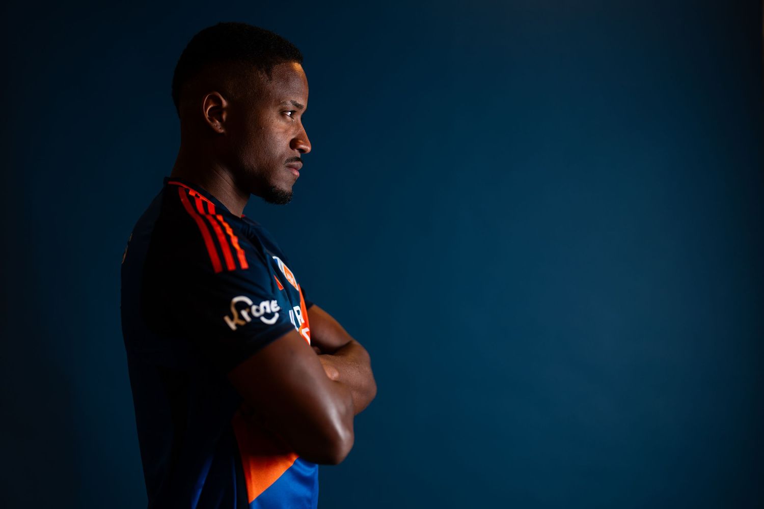 Side-profile portrait of a person wearing a blue and orange FC Cincinnati jersey with arms crossed against a blue background.