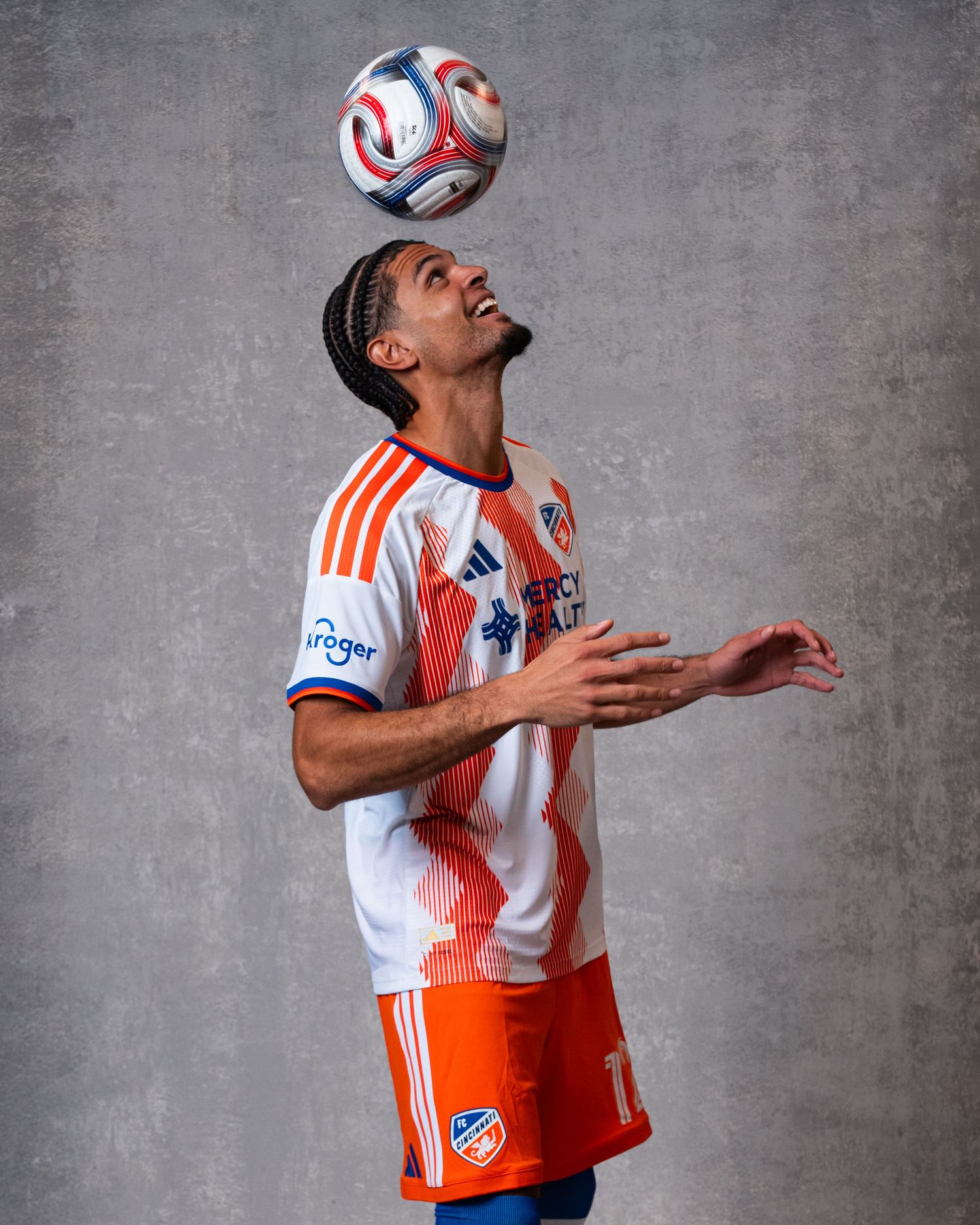 Person balancing a soccer ball above their forehead while wearing a white and orange FC Cincinnati uniform against a gray studio backdrop.
