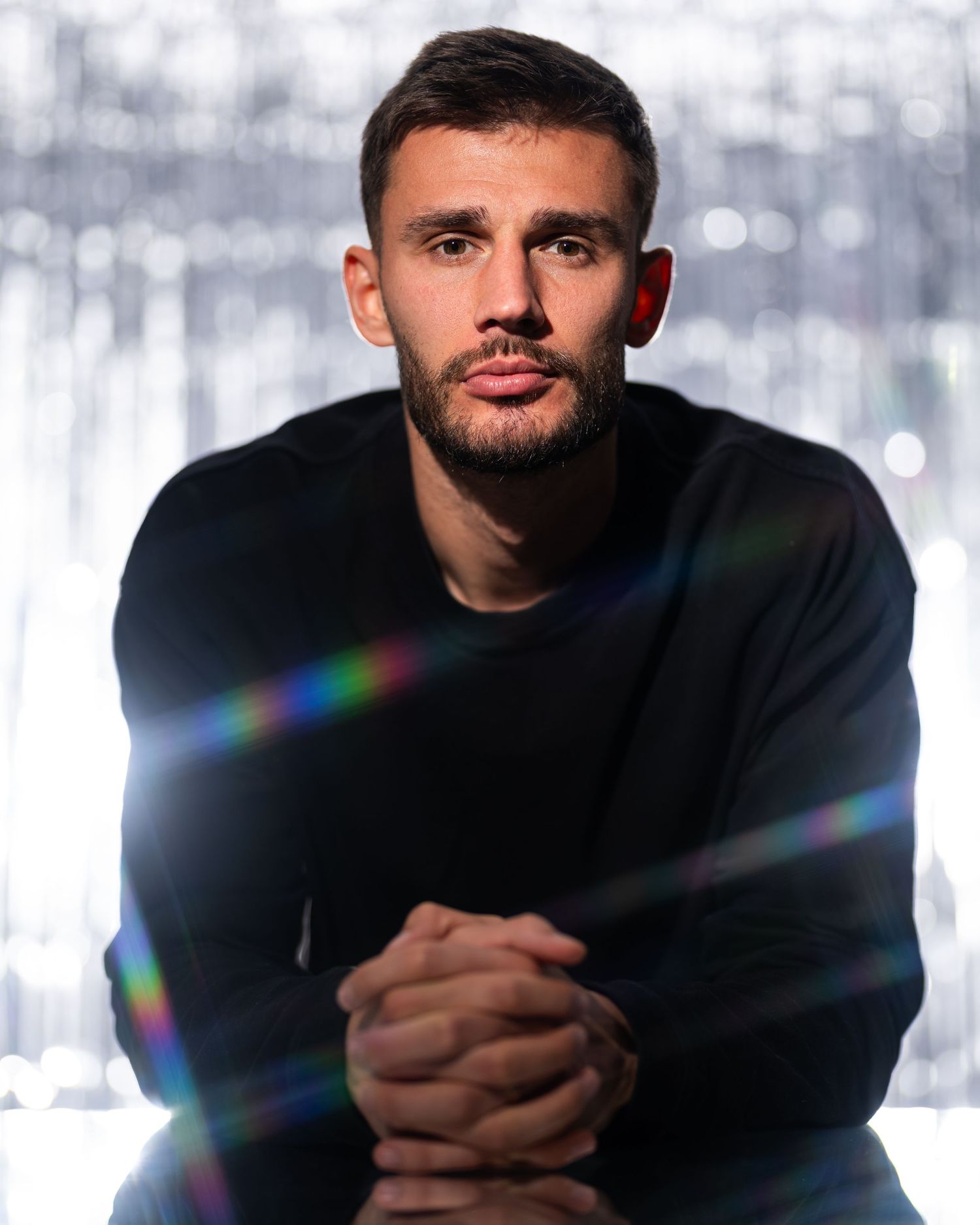 Studio portrait of a soccer player seated with hands clasped against a bright reflective backdrop.