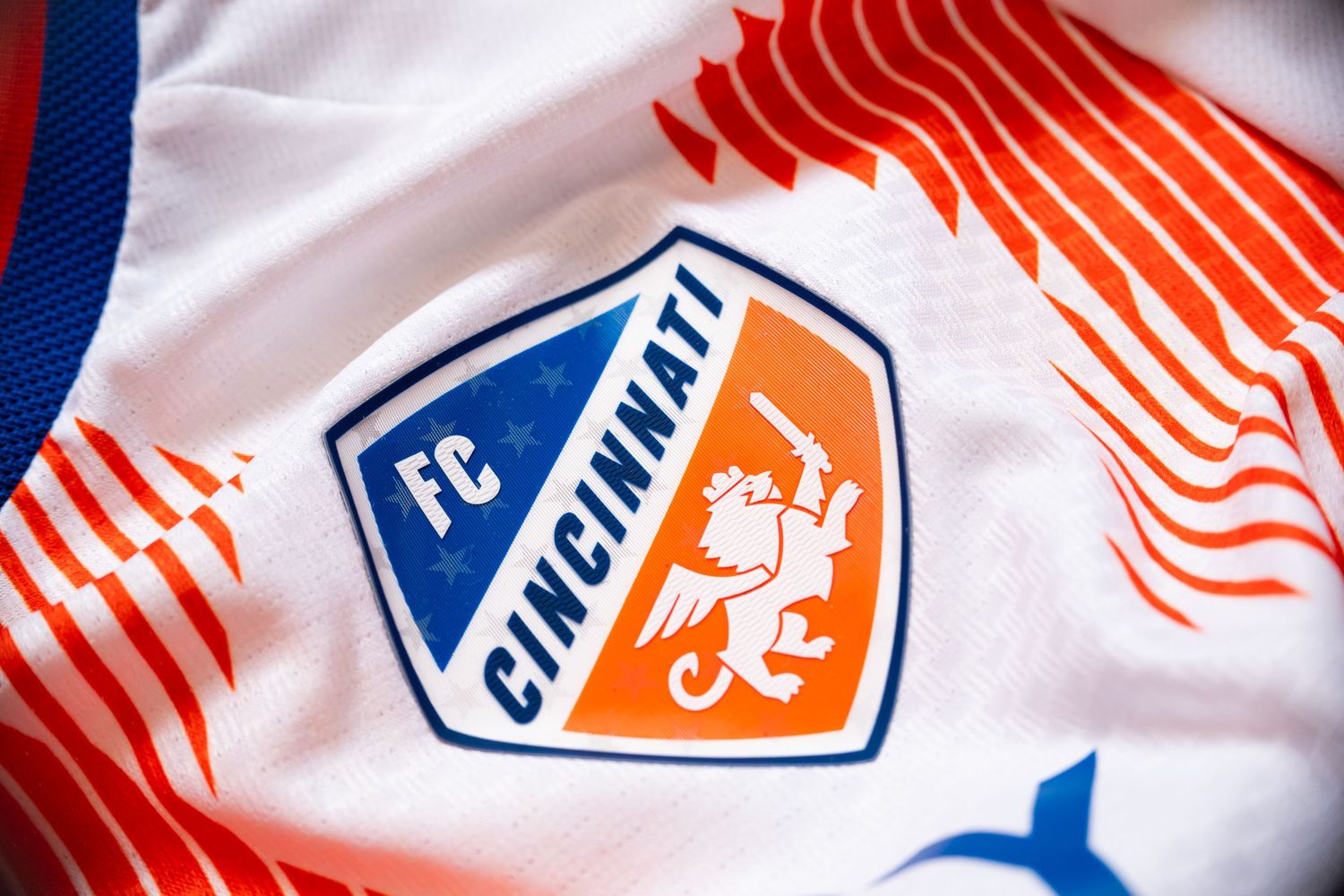 Close-up of the FC Cincinnati crest on a white jersey with orange graphic striping and blue trim.