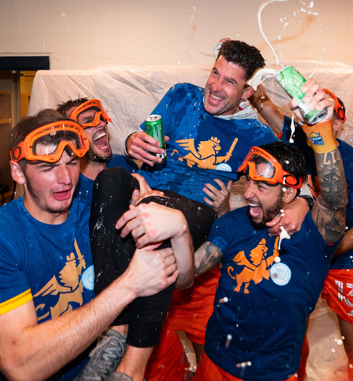 Soccer players wearing protective goggles celebrate in a locker room while spraying beverages after a victory.