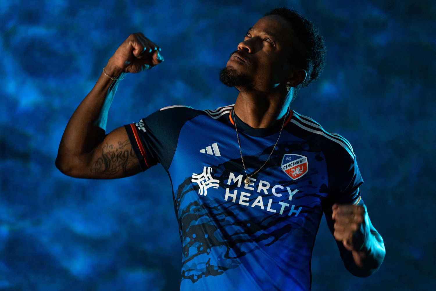 Person wearing a blue FC Cincinnati jersey posing with raised arms against a blue textured backdrop.