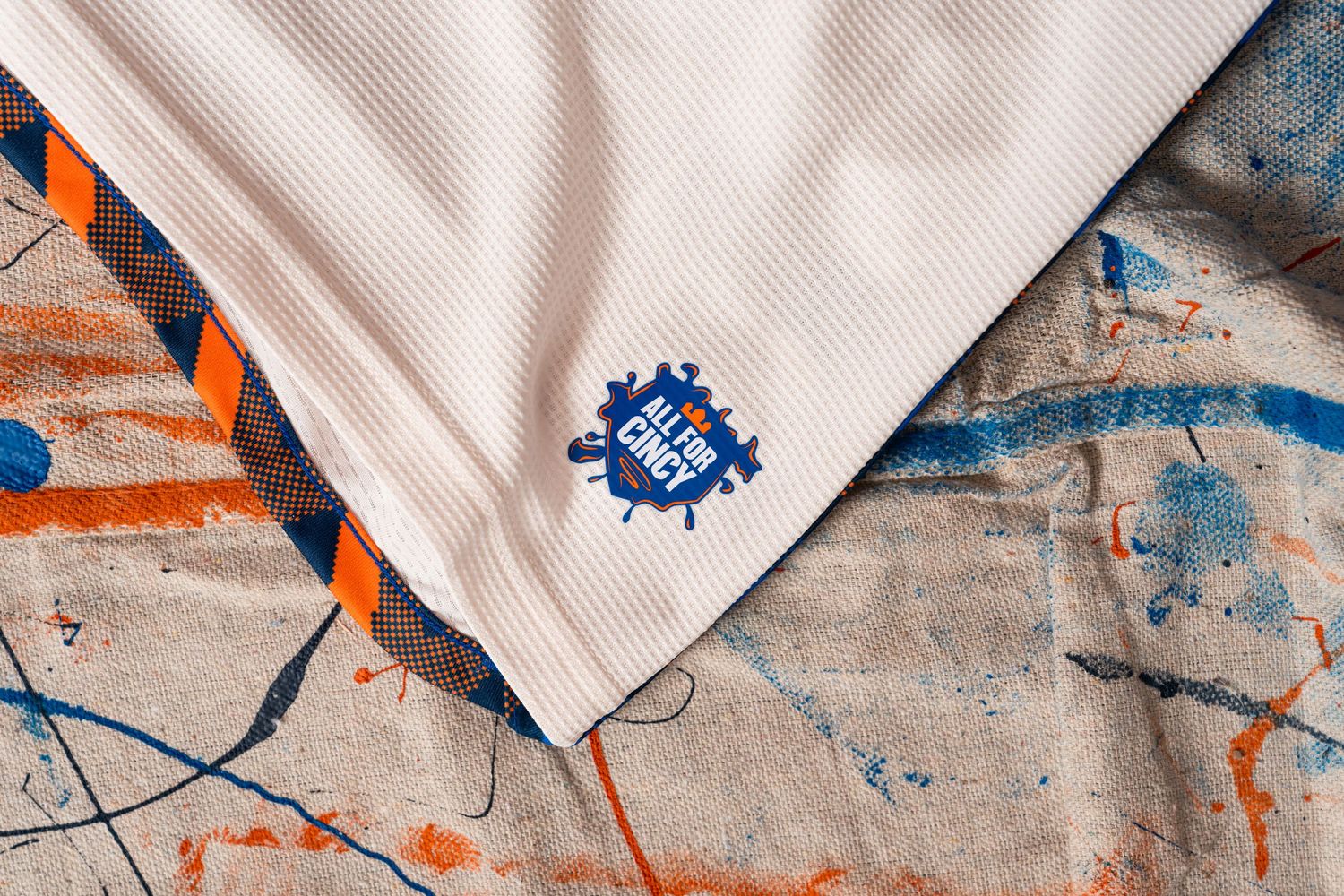 Close-up detail of a white jersey hem featuring an “All For Cincy” graphic above a paint-splattered canvas drop cloth.