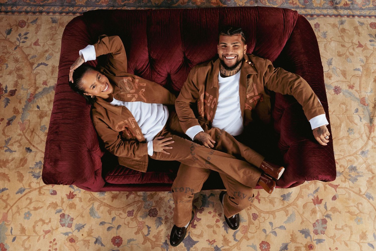 Overhead view of two people reclining on a red velvet sofa while wearing matching brown FC Cincinnati apparel.
