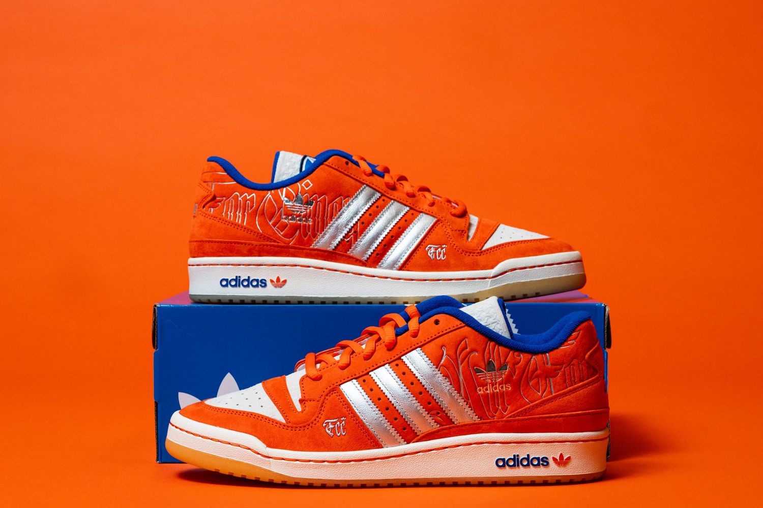 Pair of orange Adidas sneakers displayed on a blue Adidas shoebox against an orange background, showing silver stripes and custom graphics.