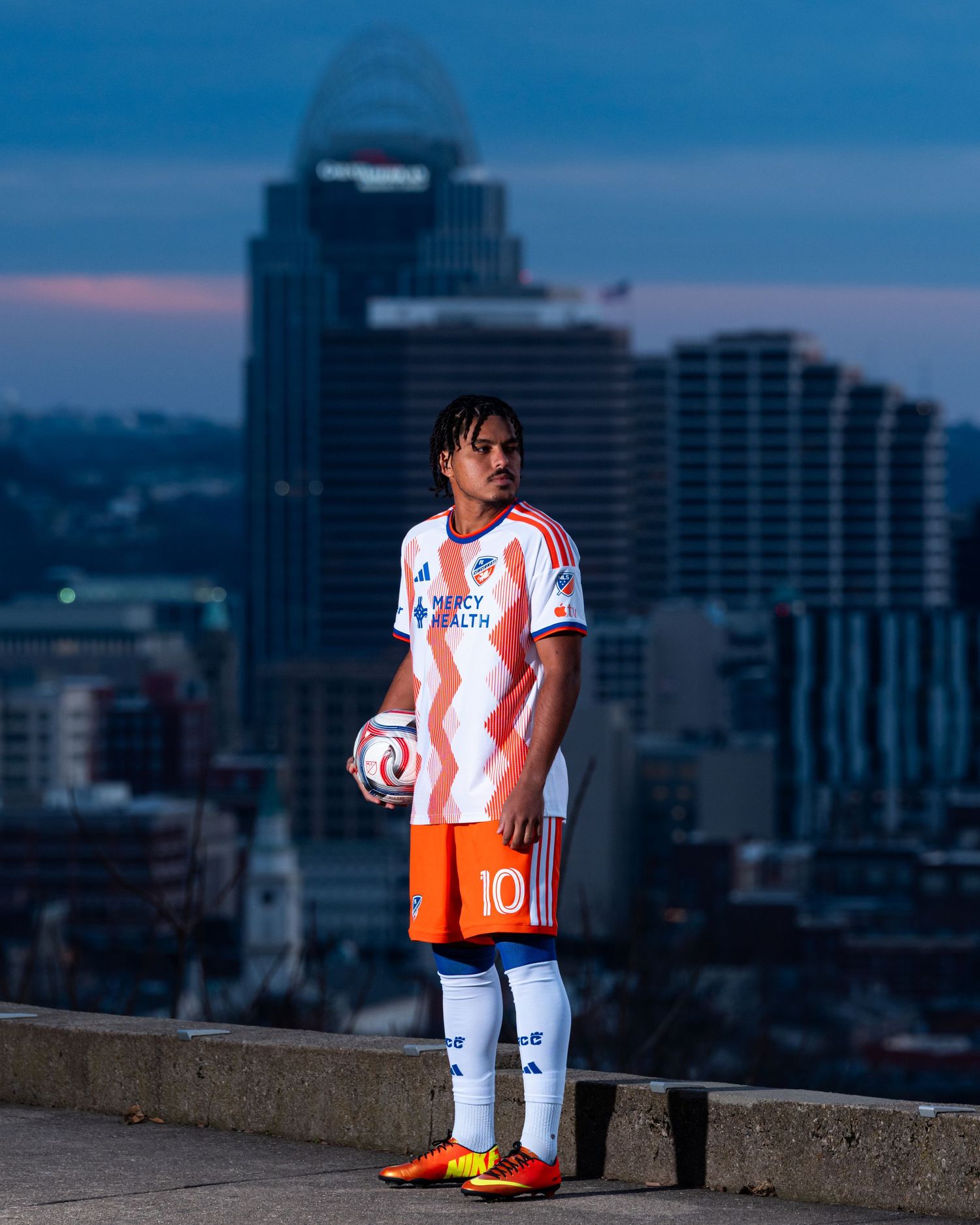 Person holding a soccer ball while wearing a white and orange FC Cincinnati uniform on an overlook with the Cincinnati skyline.