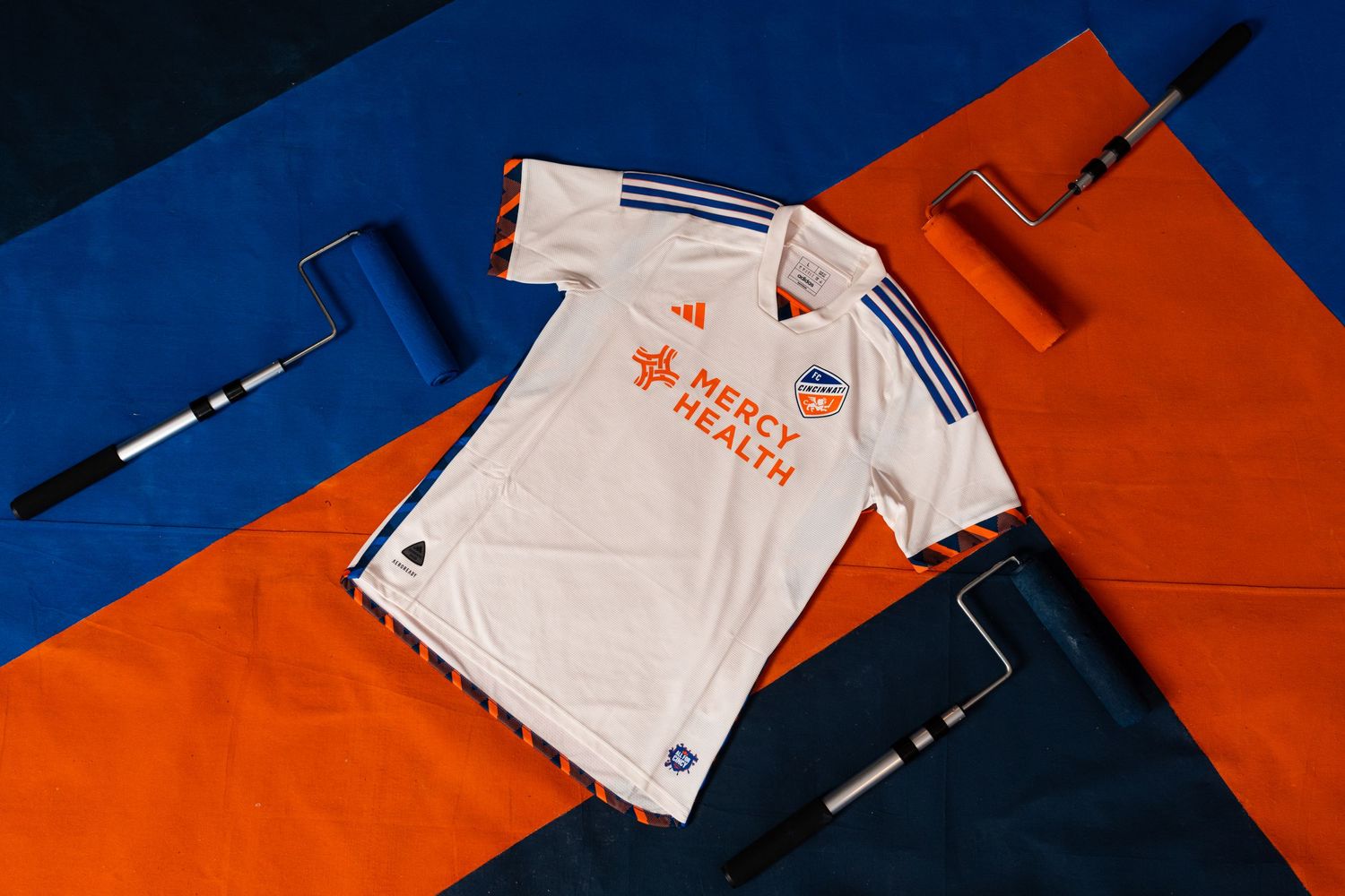 Flat-lay view of a white FC Cincinnati jersey surrounded by paint rollers on a blue and orange geometric background.