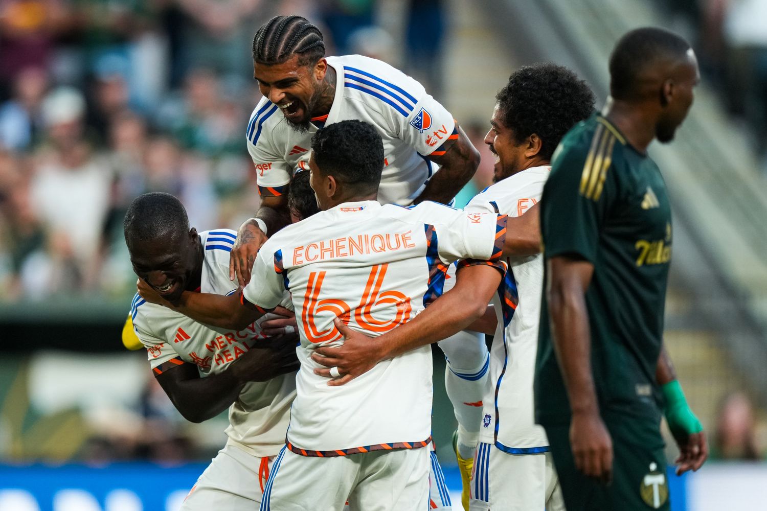 FC Cincinnati players in white uniforms celebrate together during a match while an opposing player stands nearby.