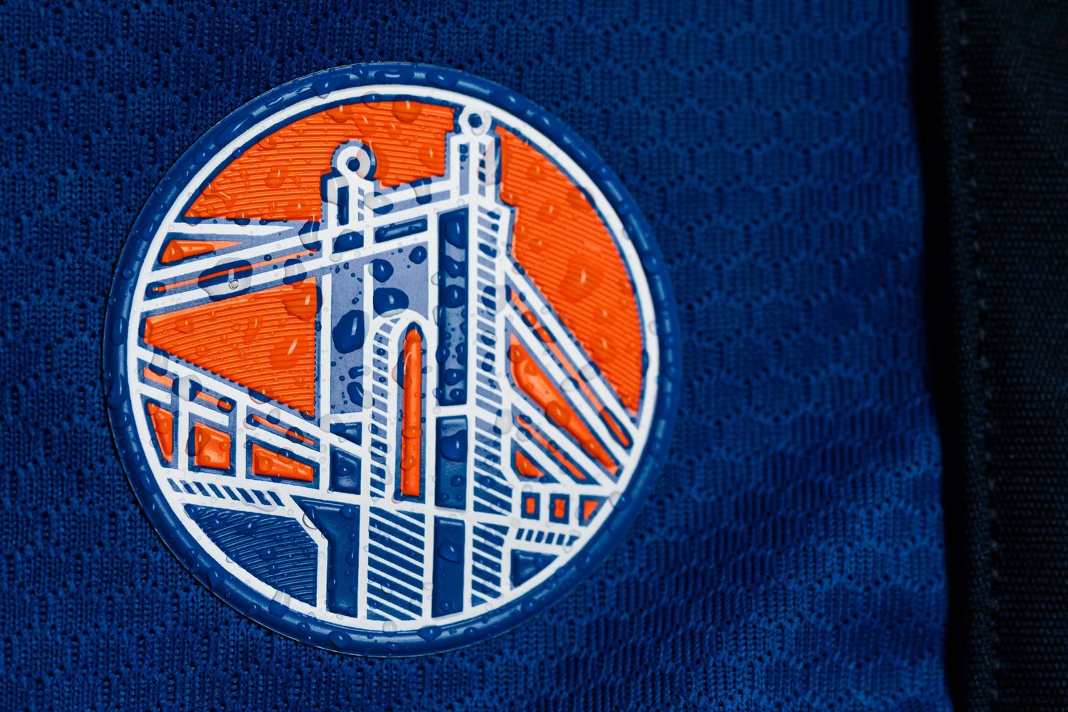 Close-up of a circular orange, blue, and white bridge-inspired emblem displayed on a blue FC Cincinnati jersey with water droplets visible.