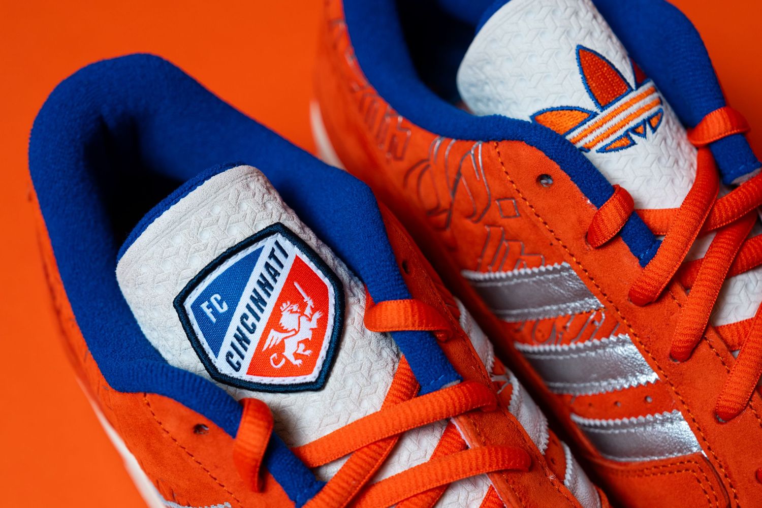 Close-up view of two orange Adidas sneakers with blue lining and orange laces, showing an FC Cincinnati crest on one tongue.