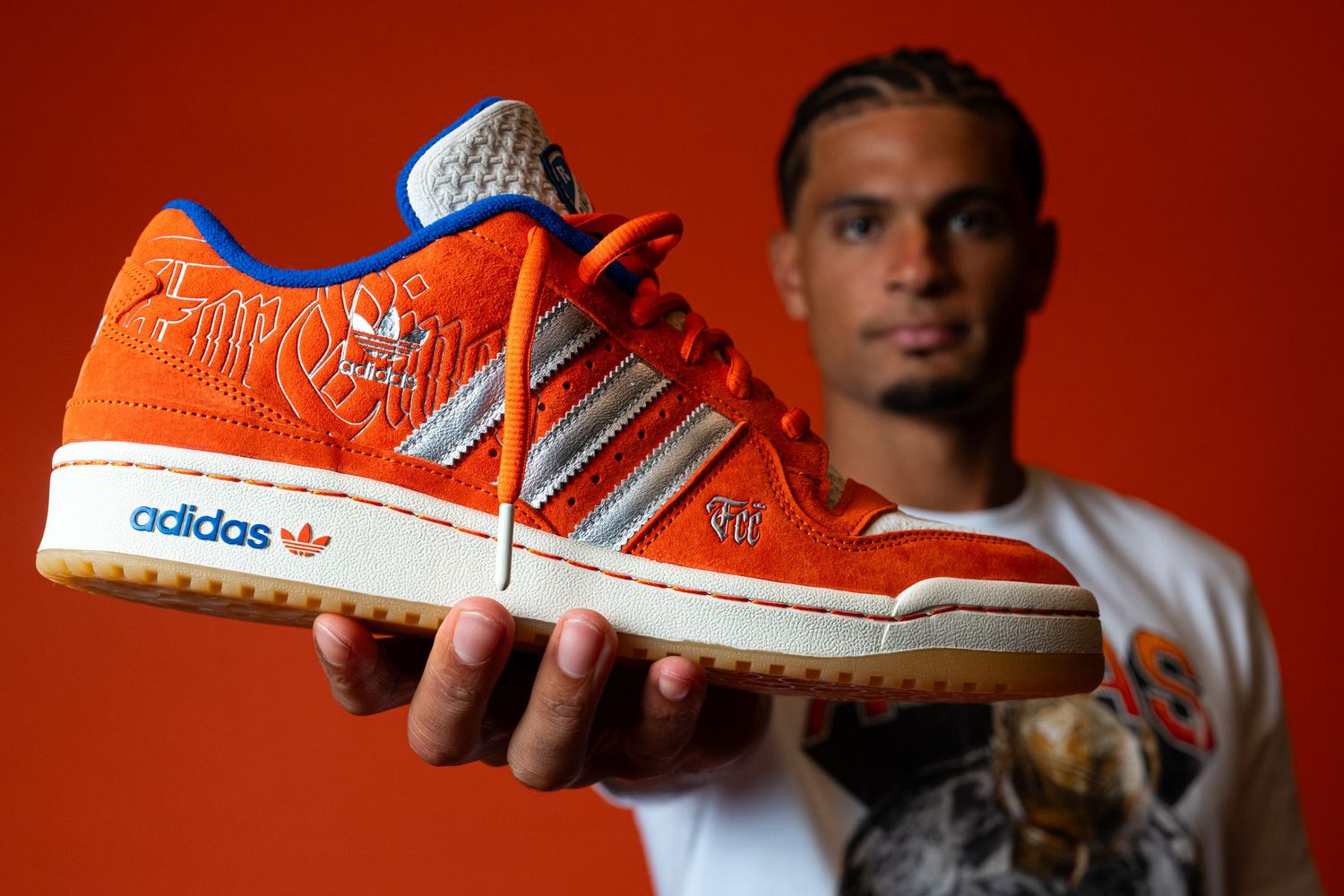 Person holding an orange Adidas sneaker toward the camera, highlighting the side profile, silver stripes, and Adidas branding.