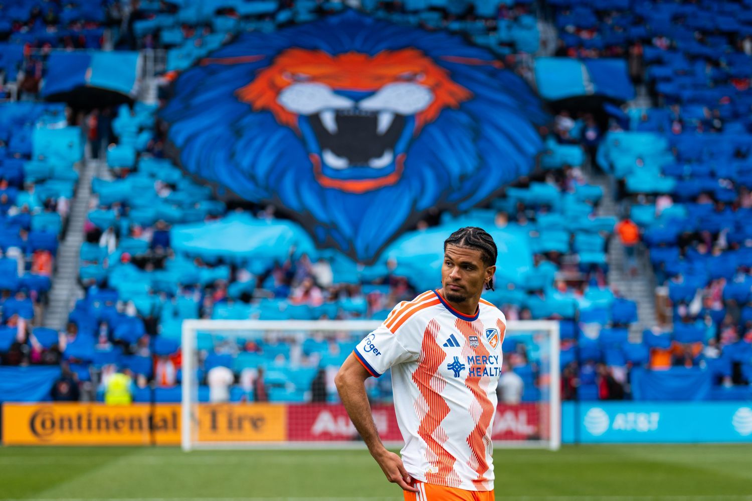 Soccer player stands on the field with a large lion tifo displayed by supporters in the stadium behind.