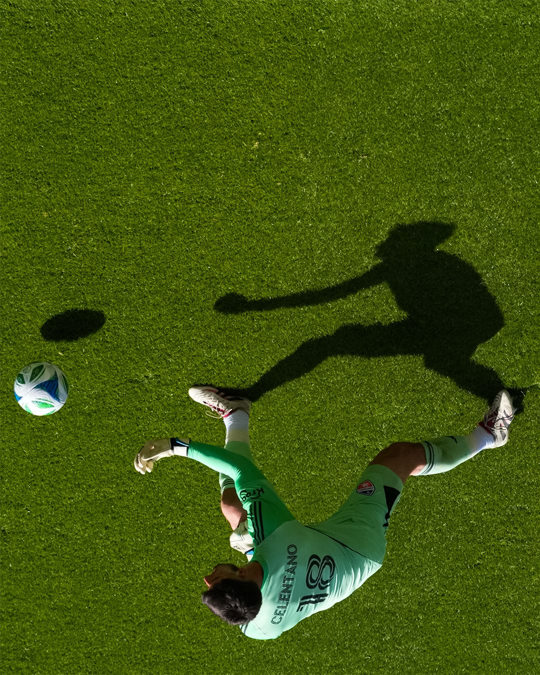 Overhead view of a goalkeeper and shadow positioned near a soccer ball on the field.