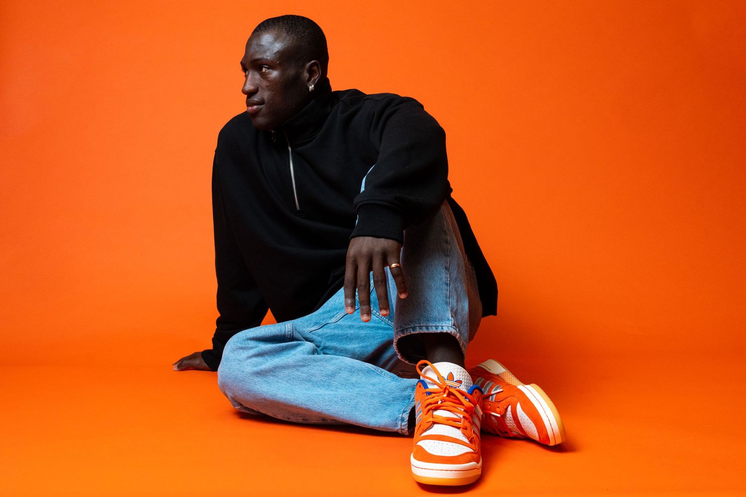 Person seated on an orange studio floor wearing orange Adidas sneakers, blue jeans, and a dark pullover.