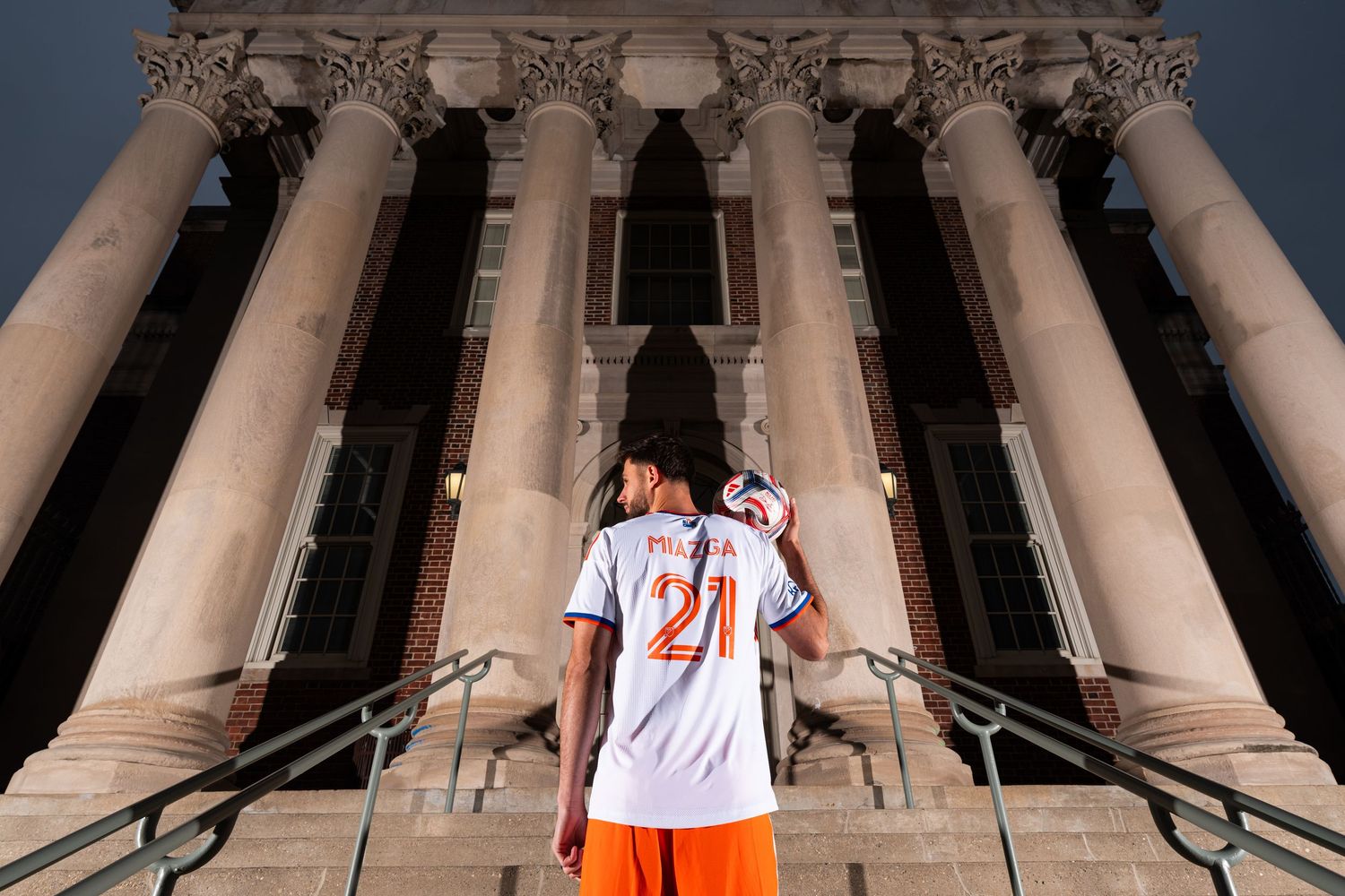 Rear view of a person holding a soccer ball while standing on steps in front of a building with tall stone columns.