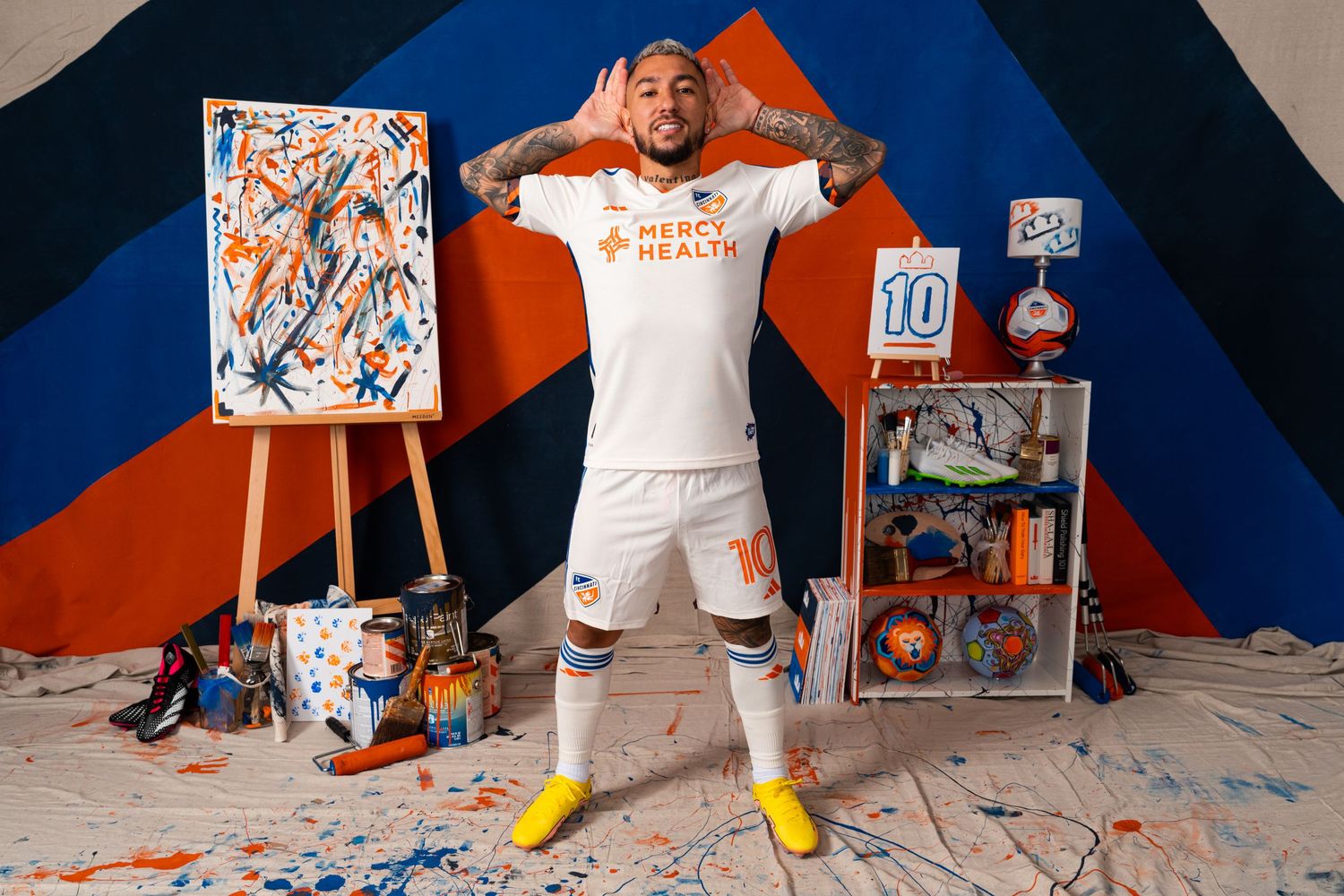 Soccer player in a white FC Cincinnati uniform posing with both hands raised beside their head in front of a painted studio.
