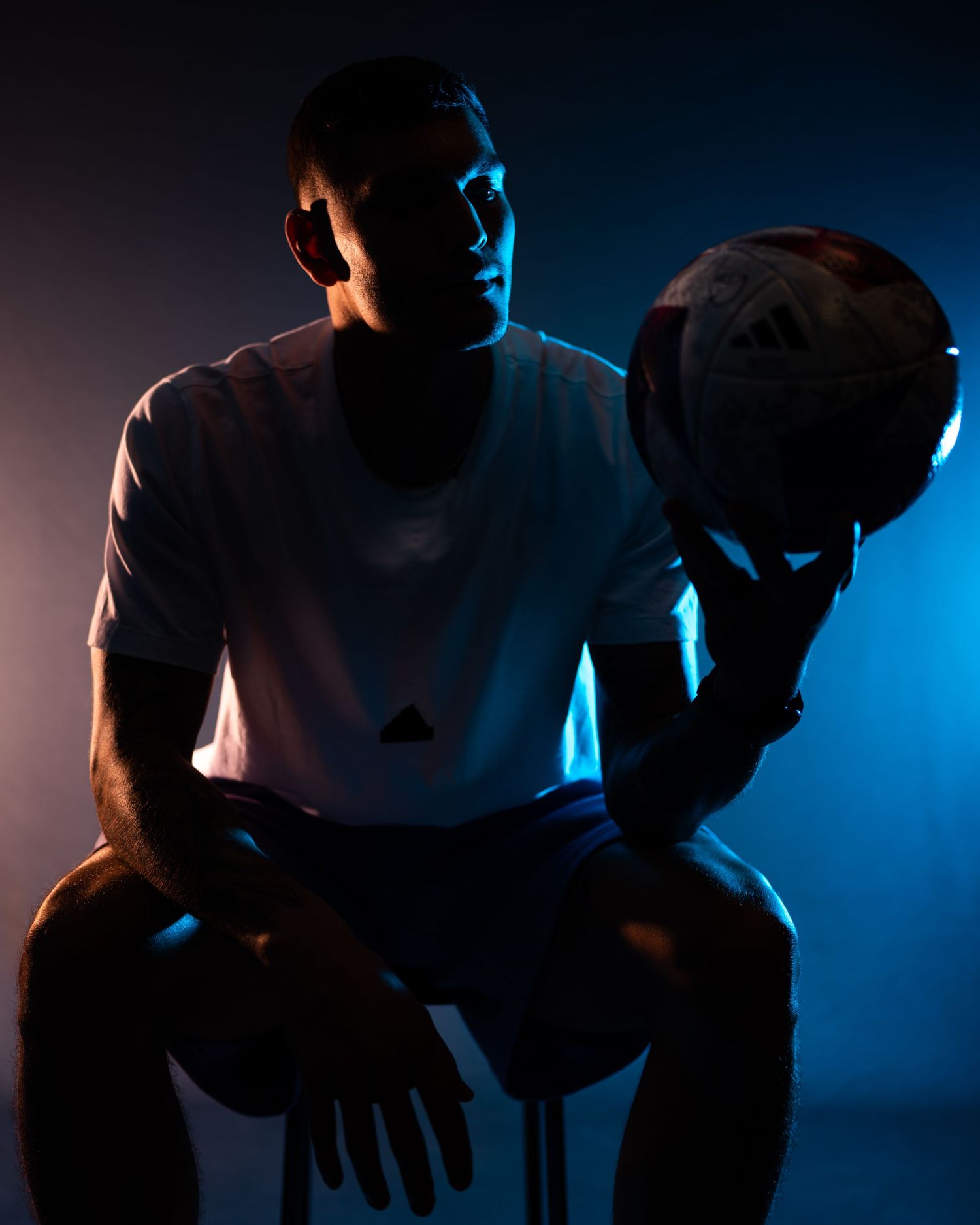 Soccer player seated in a studio holding a soccer ball under dramatic blue and orange lighting.