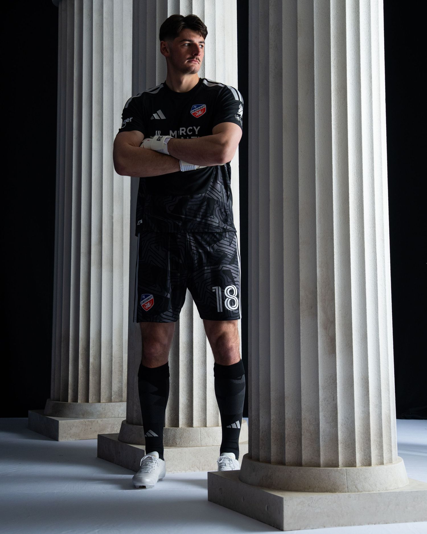 Person wearing a black FC Cincinnati goalkeeper uniform standing with arms crossed among a row of white stone columns.
