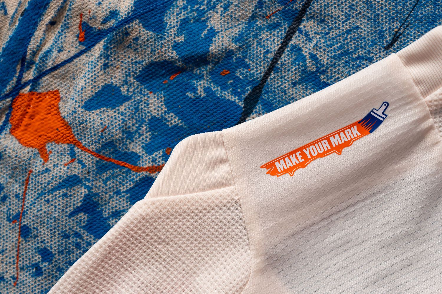 Close-up of a white jersey shoulder displaying a “Make Your Mark” graphic on a paintbrush illustration against a paint-splattered background